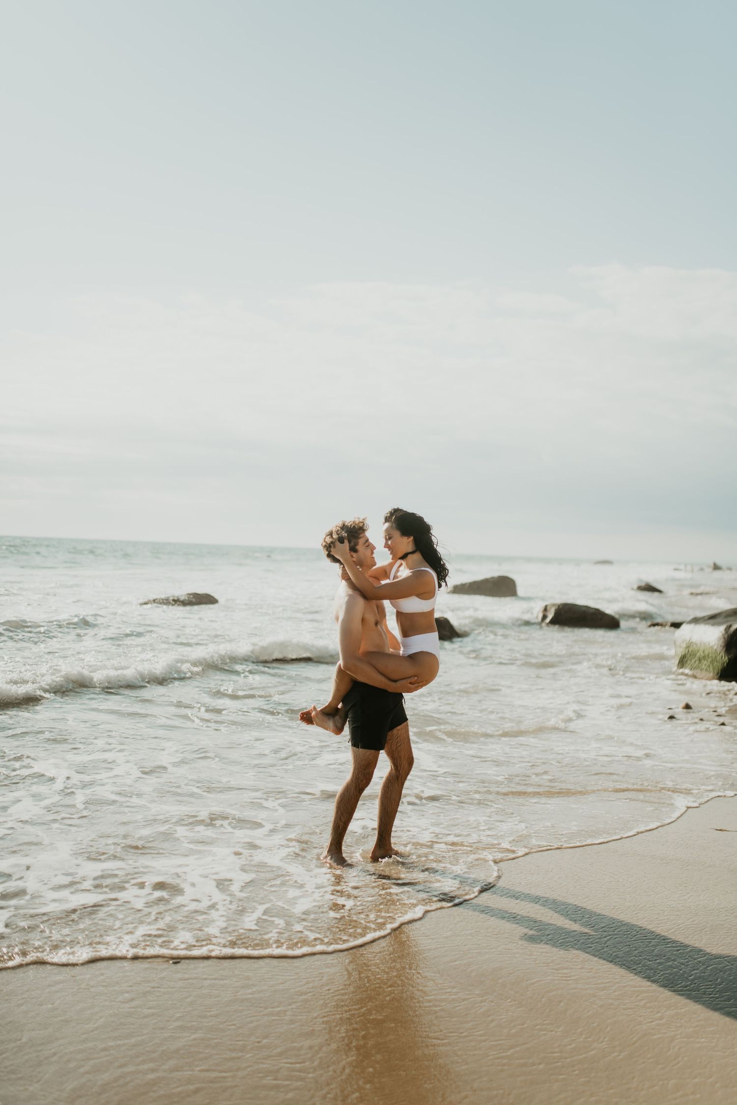Two people embrace lovingly at the beach with waves crashing on the shore and rocks in the background during sunset.