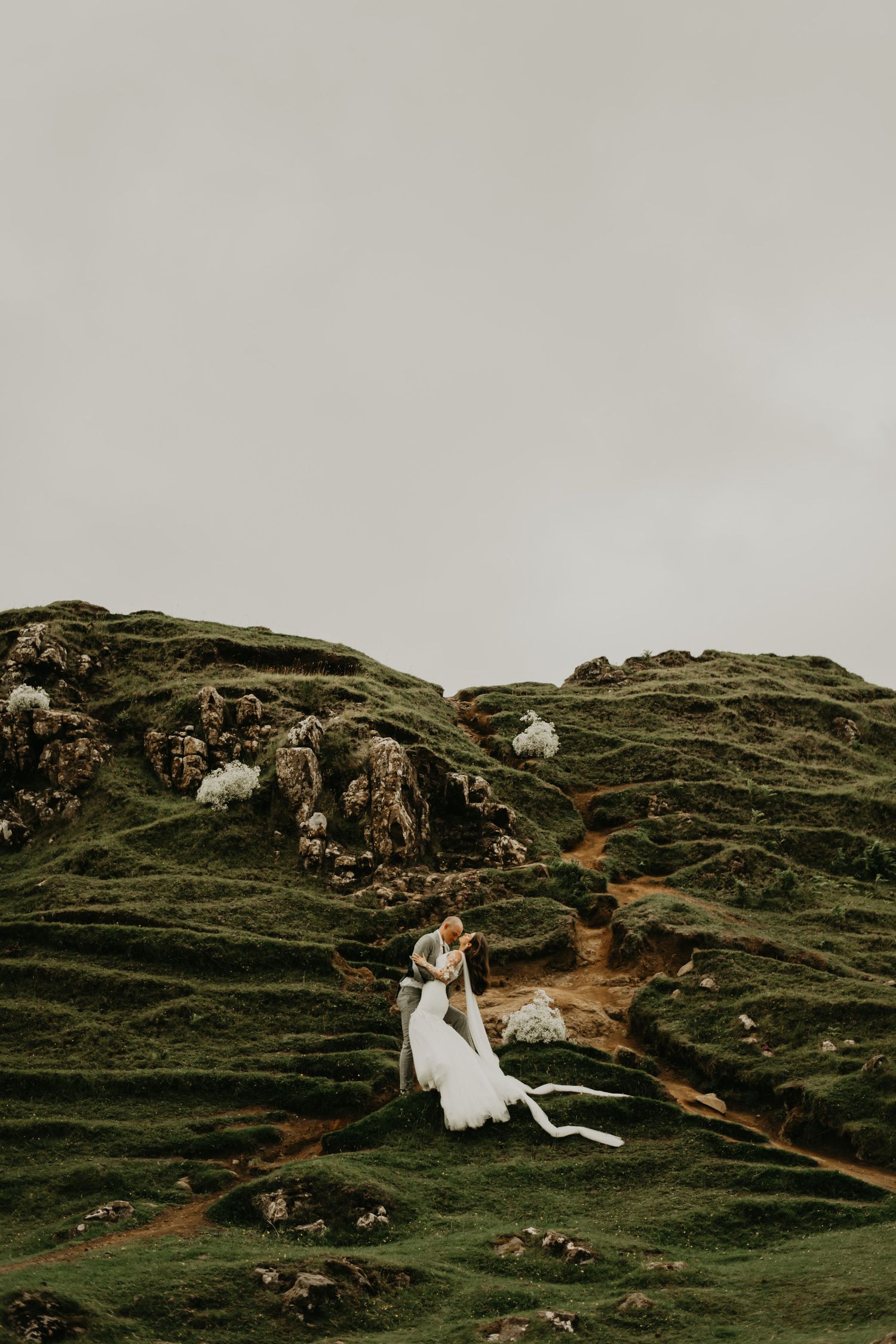 A couple in wedding attire stands on rugged green hillside with winding dirt paths in Isle of Skye Scotland.