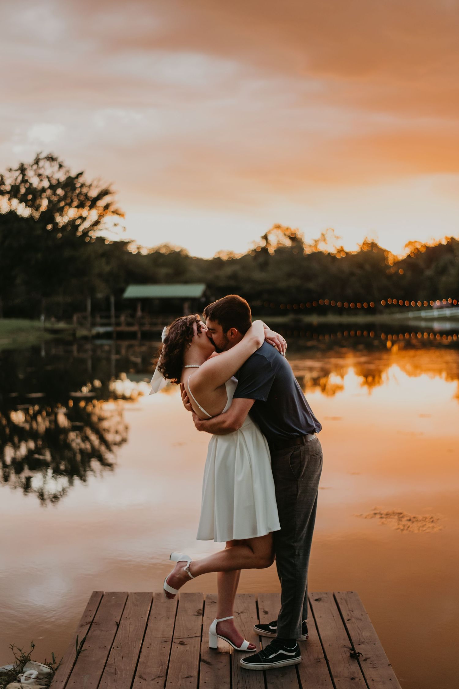 A couple embraces on a wooden dock overlooking a tranquil lake during a dramatic orange sunset.