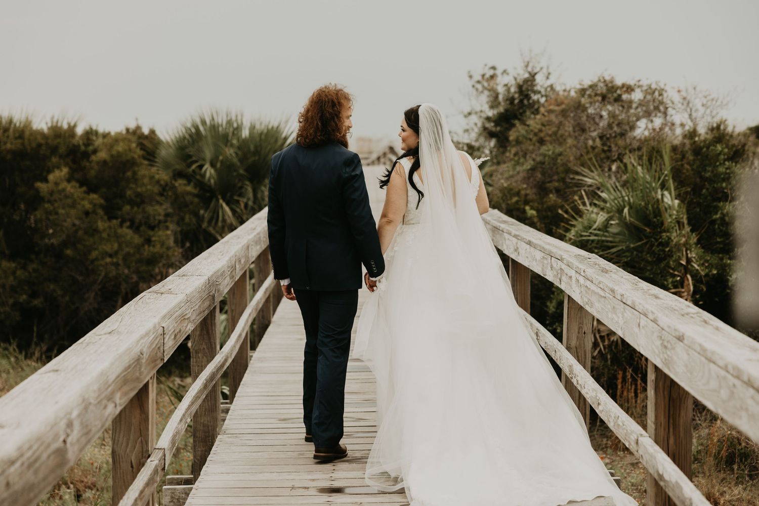 A wedding couple walks along a white wooden boardwalk surrounded by green trees.