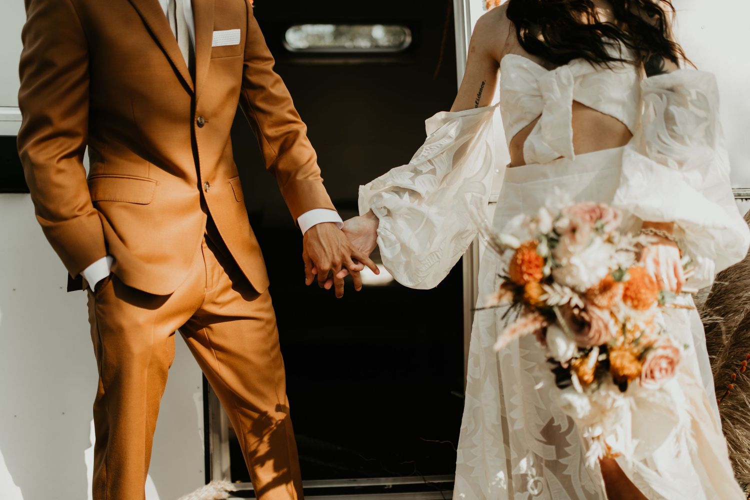 Couple in orange suit and white wedding dress holding hands in front of dark doorway in Cape Cod.