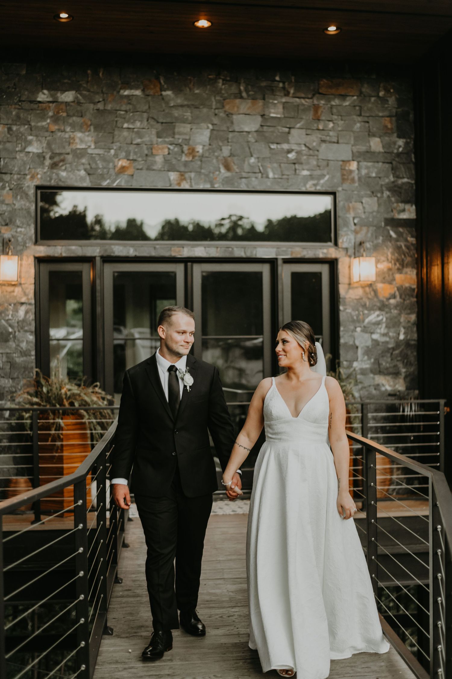 Elegant couple walks along modern stone building entrance in formal wedding attire in Asheville north Carolina.
