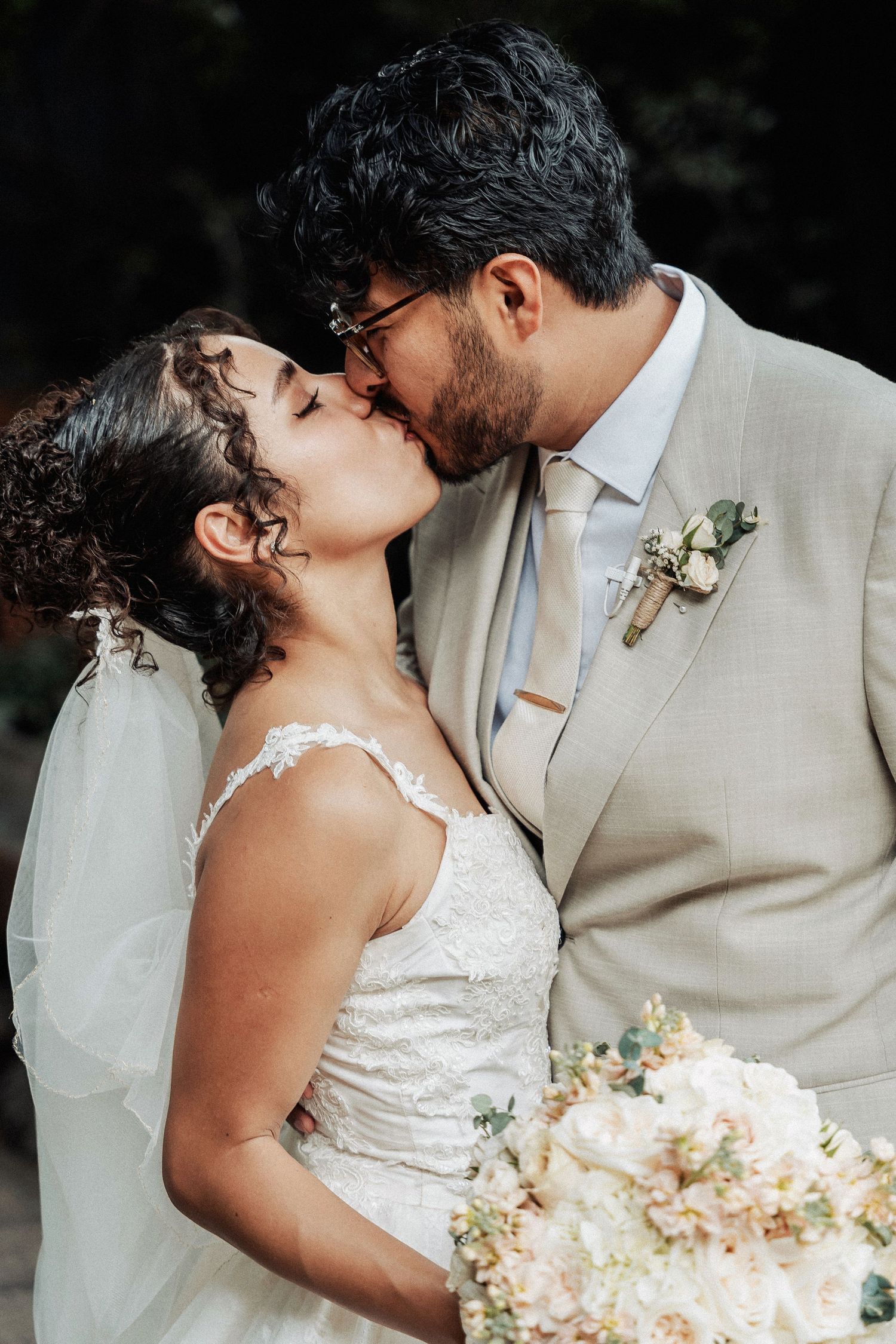 Romantic wedding photo of newlyweds sharing a kiss in beige suit and white lace dress with a soft neutral bouquet.