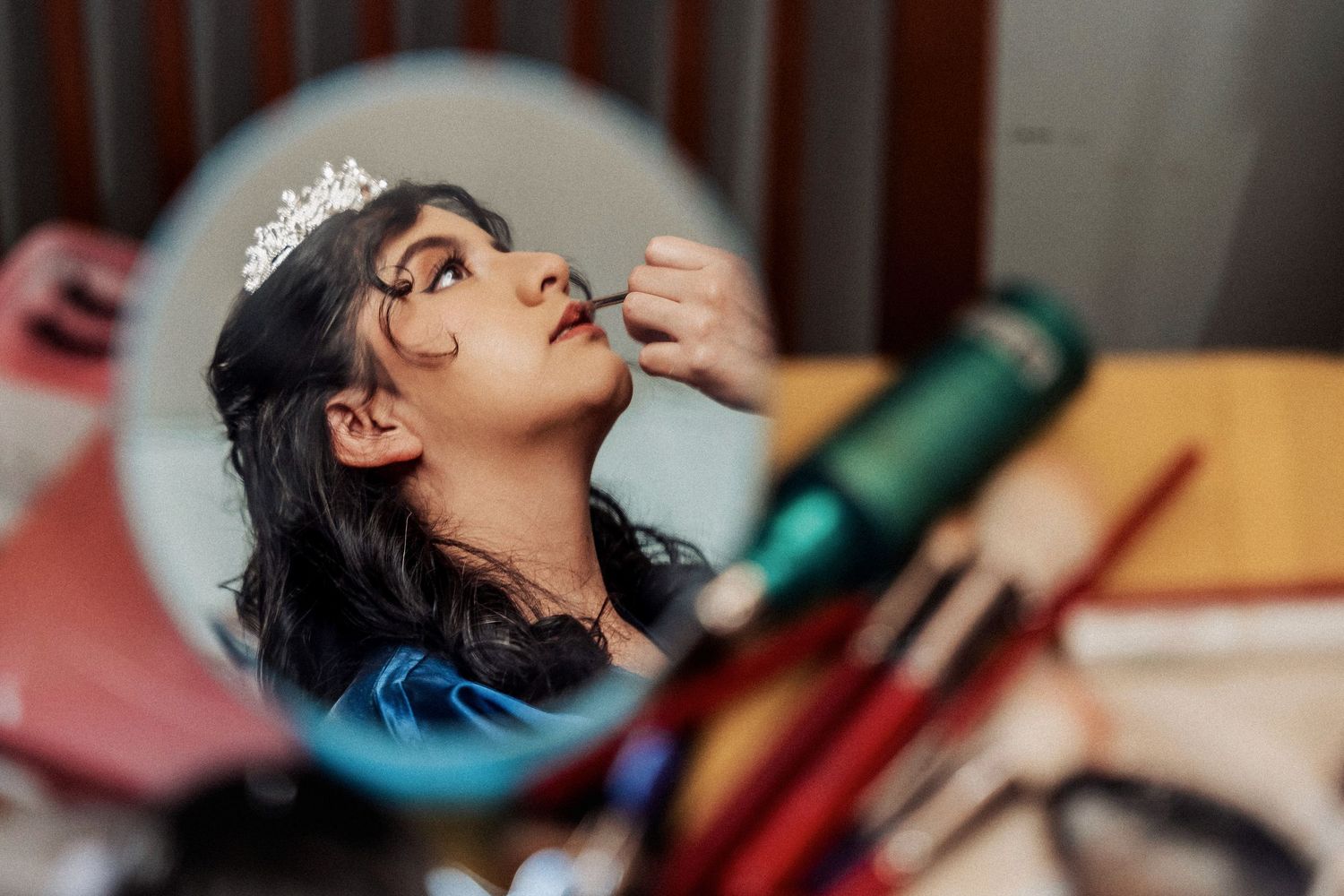 A woman wearing a tiara has her eyes closed while someone applies makeup to her face.