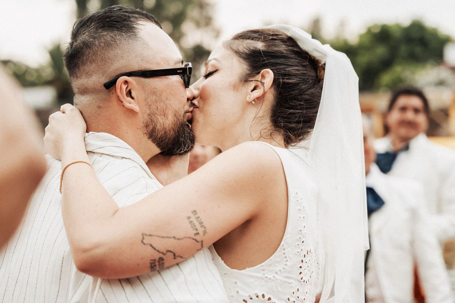 A couple shares a romantic kiss during their wedding ceremony while wearing white attire and surrounded by guests outdoors.