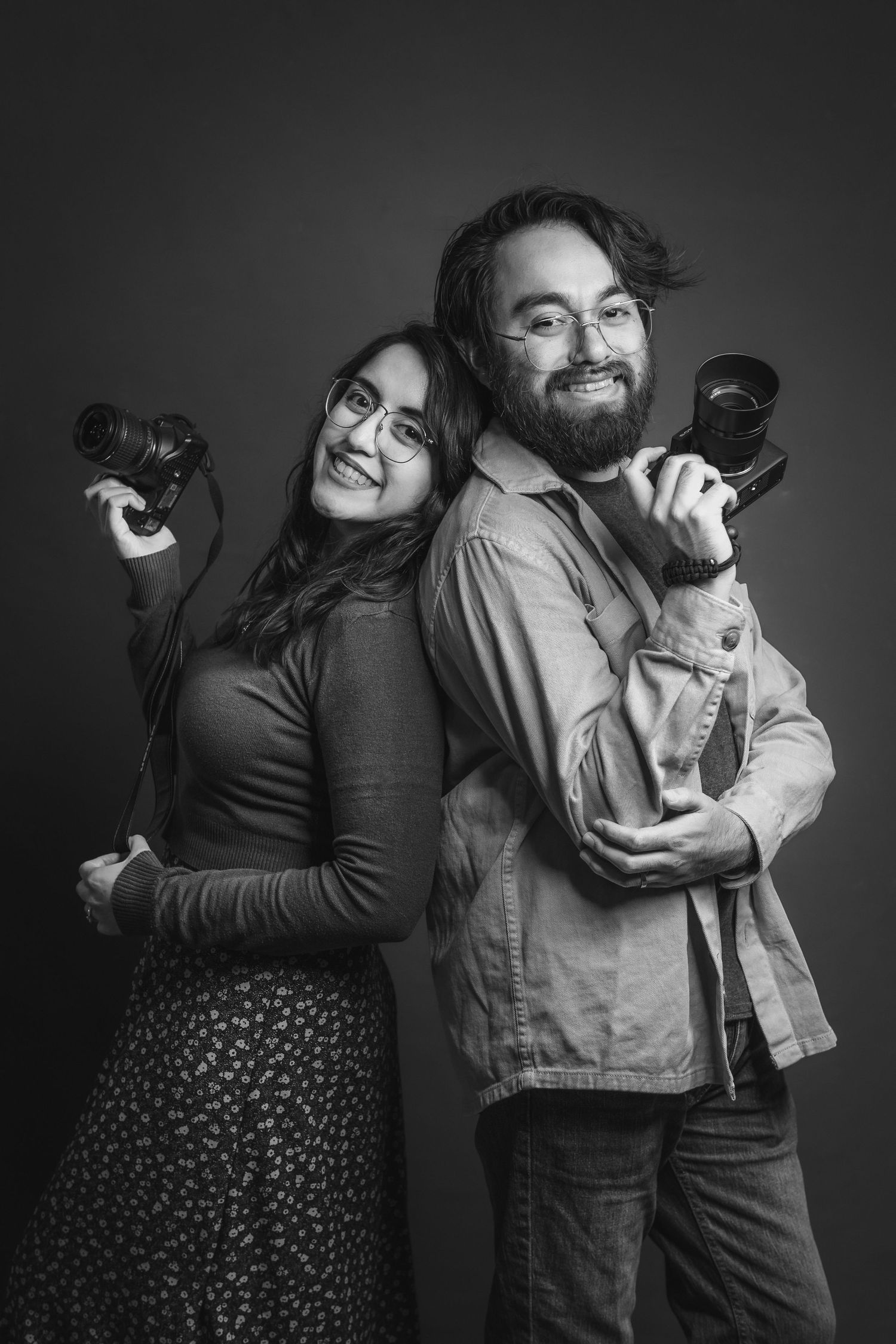 Two photographers posing back to back with cameras in a dramatic black and white studio portrait.