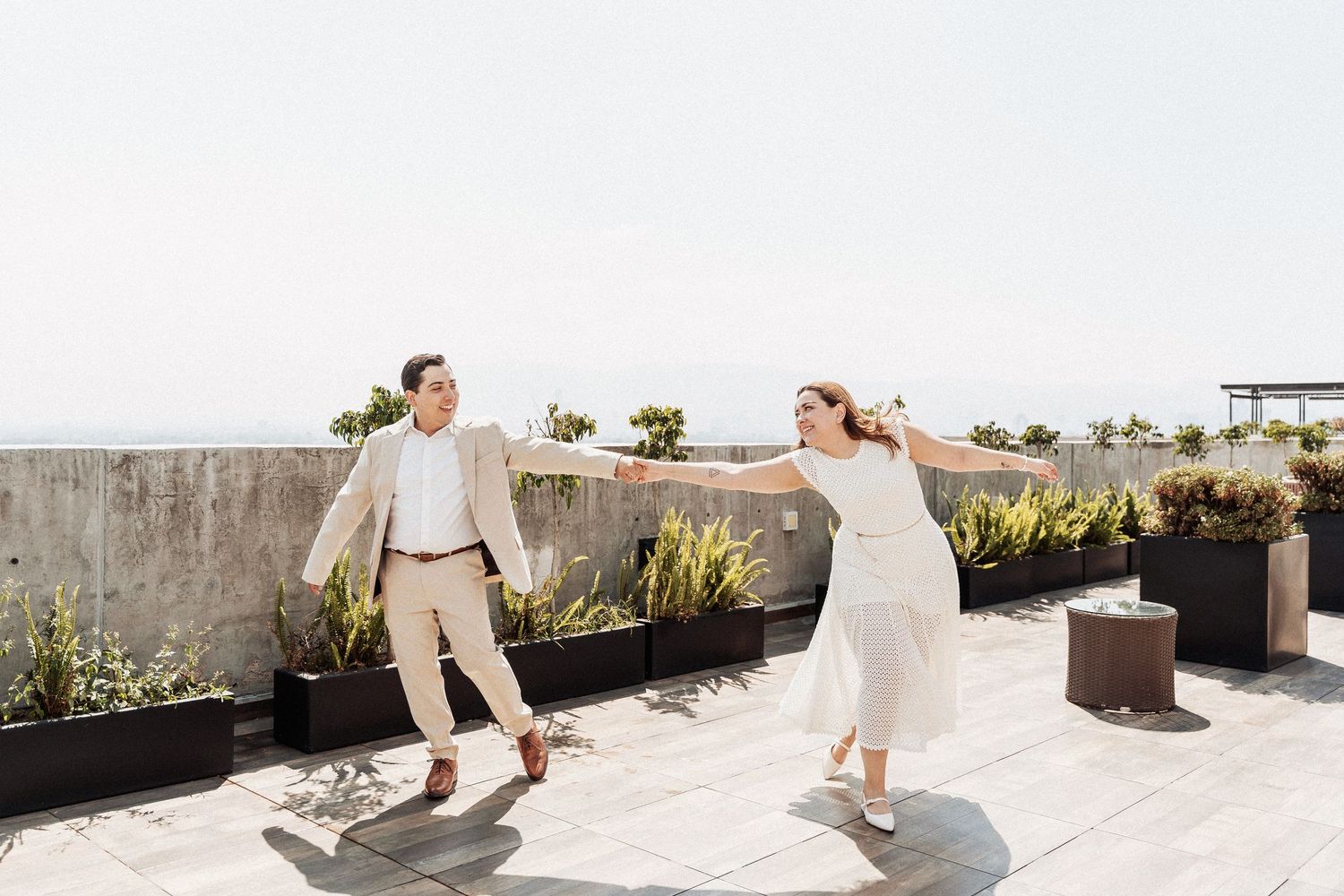 A couple dances joyfully on a rooftop terrace with plants and city views in the background.