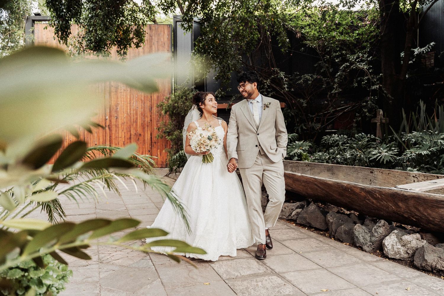 Couple walks together on stone pathway in garden setting wearing light colored wedding attire.