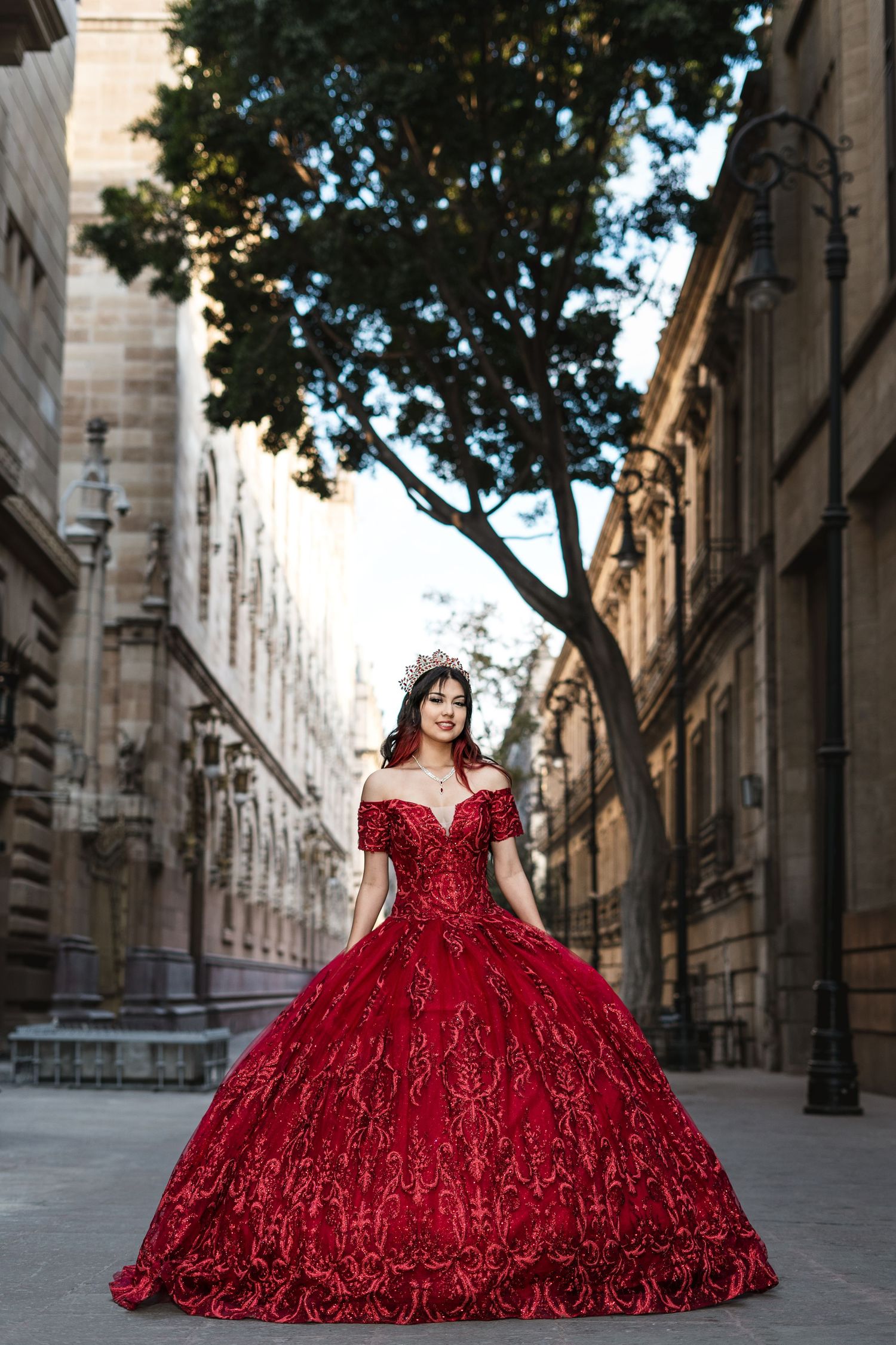 Person in an ornate red ballgown posing in front of historic stone buildings on a cobblestone street.