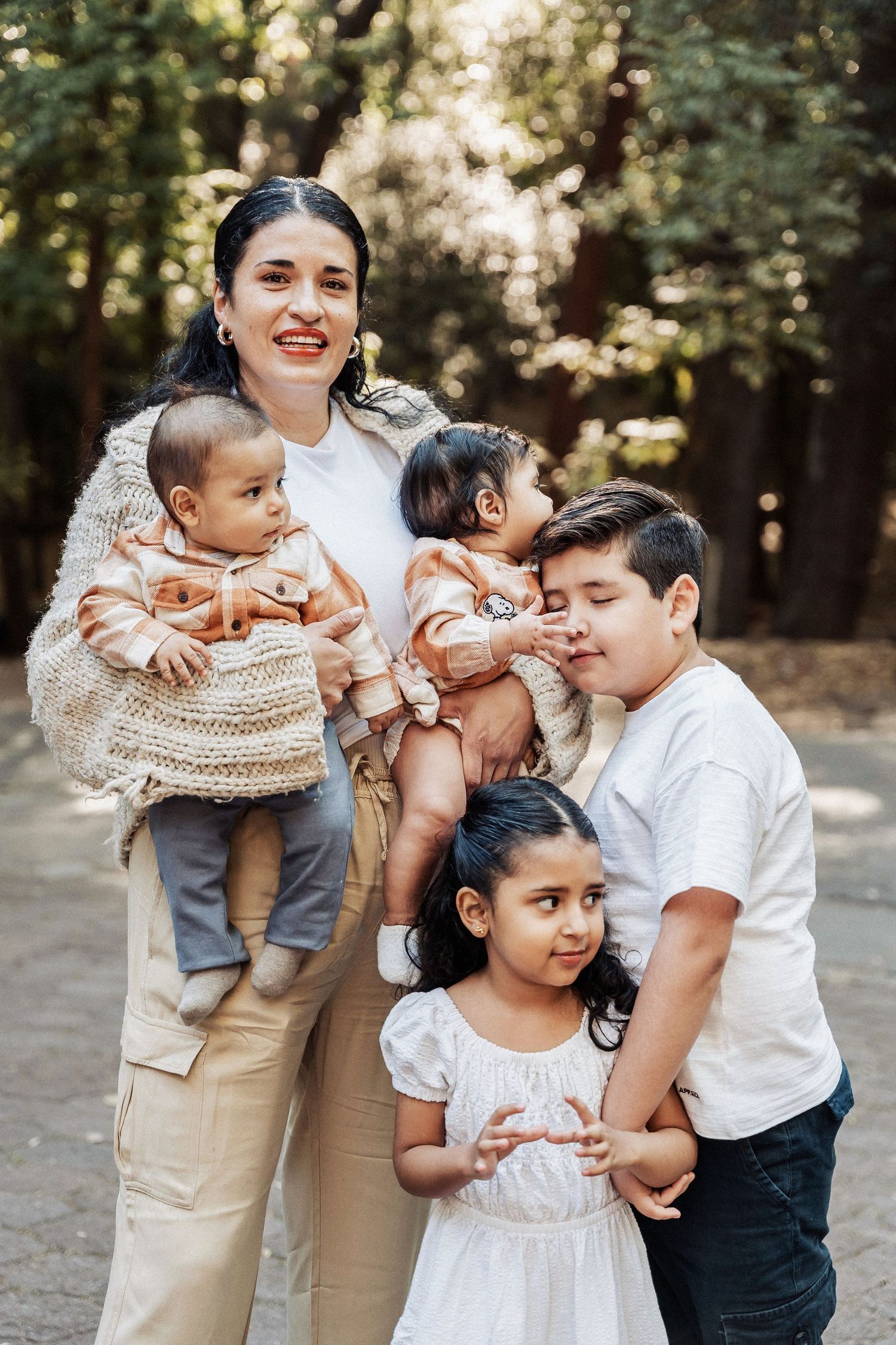 Family wearing white and beige outfits poses together in a wooded outdoor setting during a casual photo session.