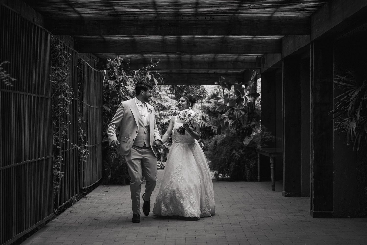 A couple walks together down a covered walkway lined with greenery in an elegant black and white wedding photo.