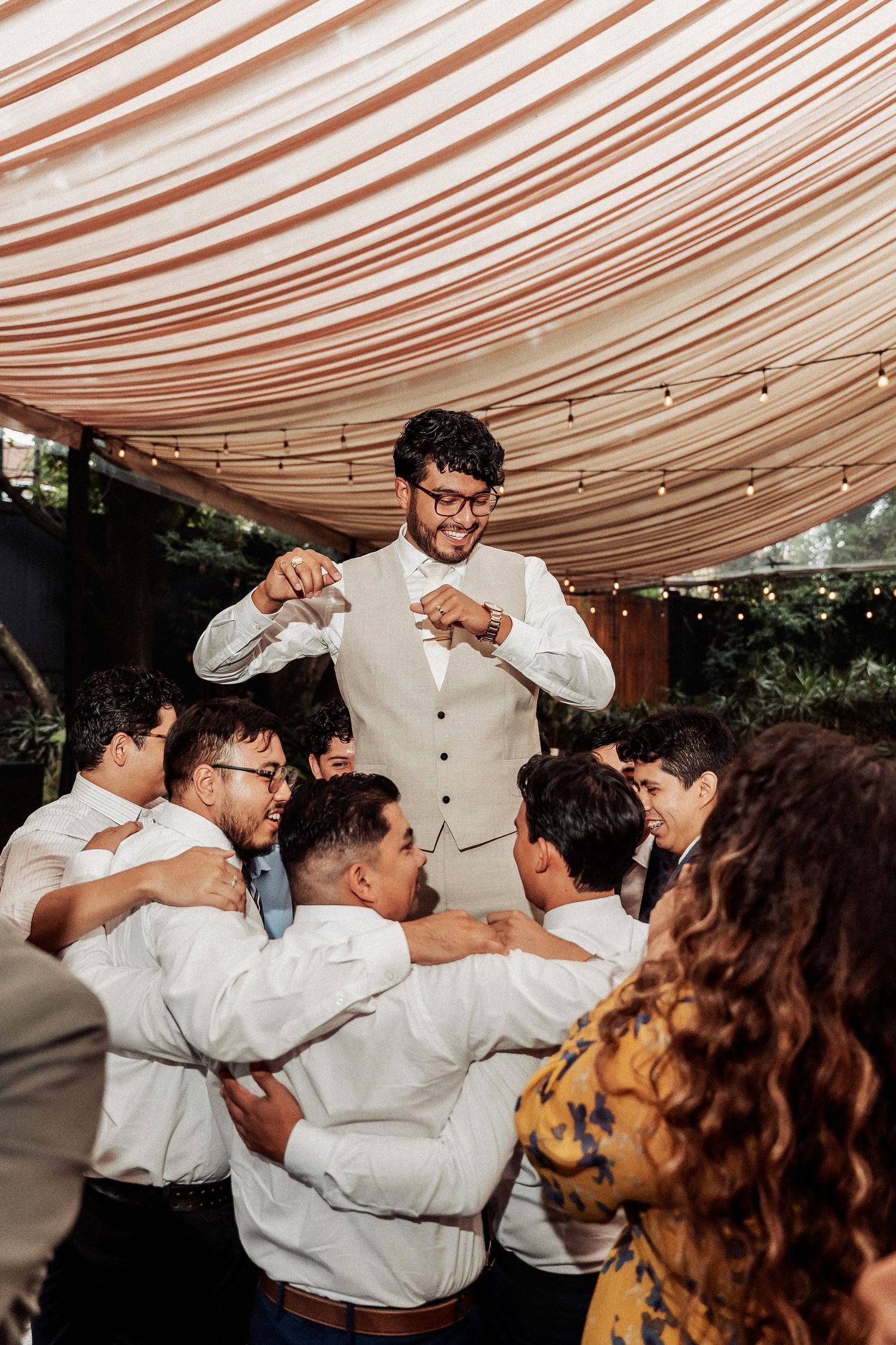 Group of guests in white shirts dancing together under a striped tent canopy at a celebration.