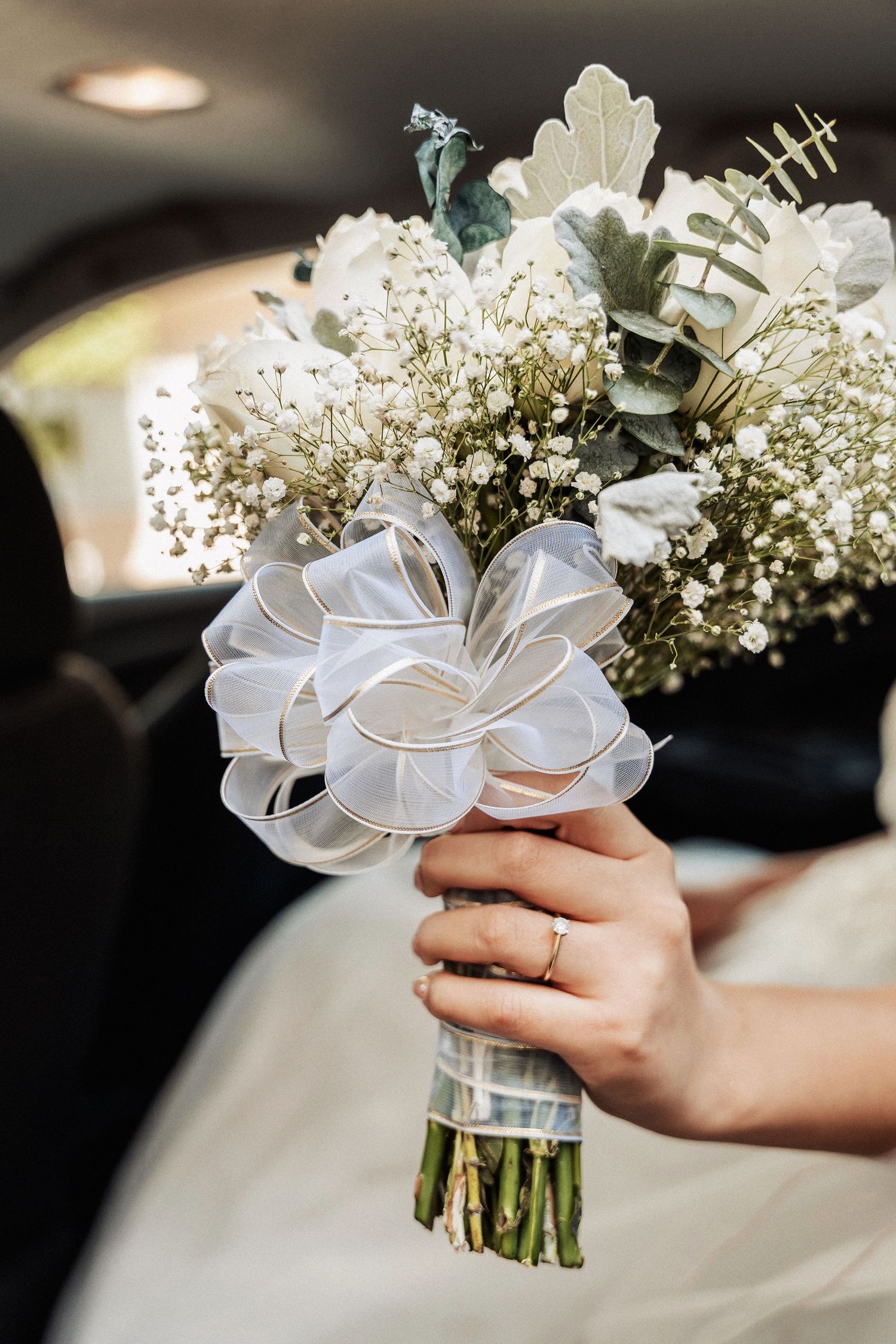 A hand wearing an engagement ring holds a delicate white bouquet with baby's breath and eucalyptus in a car.