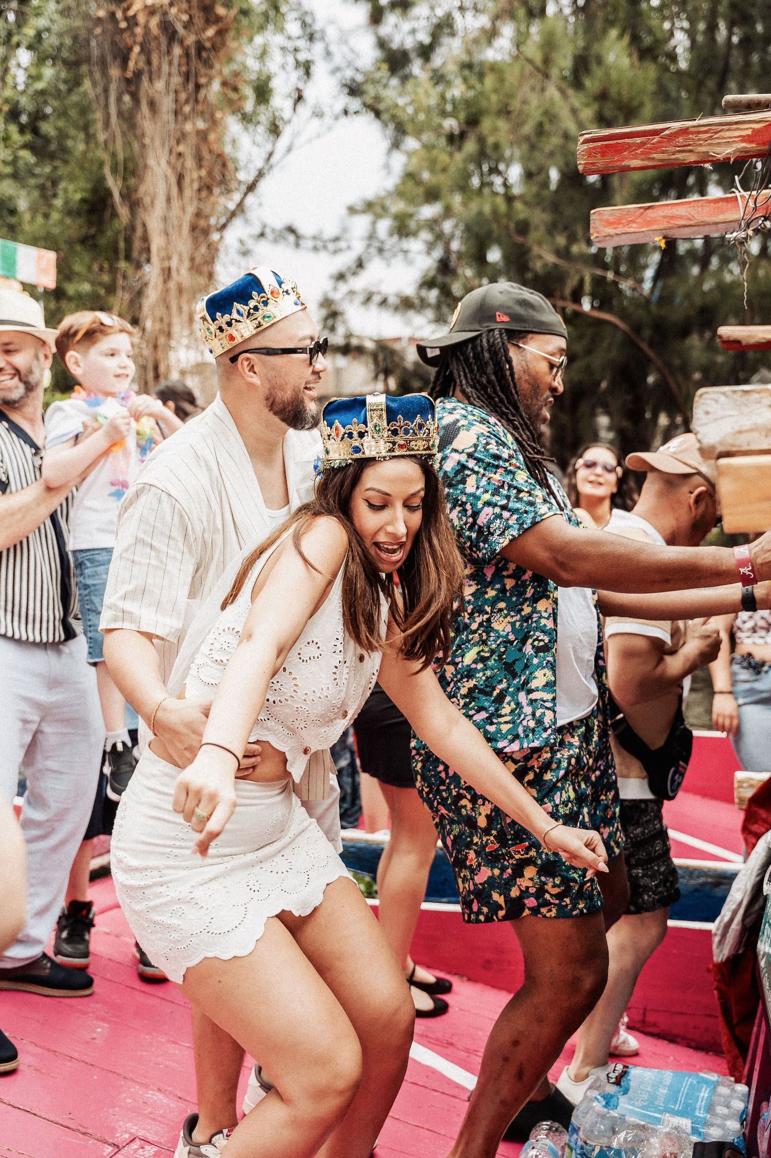 People dancing and celebrating at an outdoor party, wearing crowns and colorful outfits on a red dance floor.