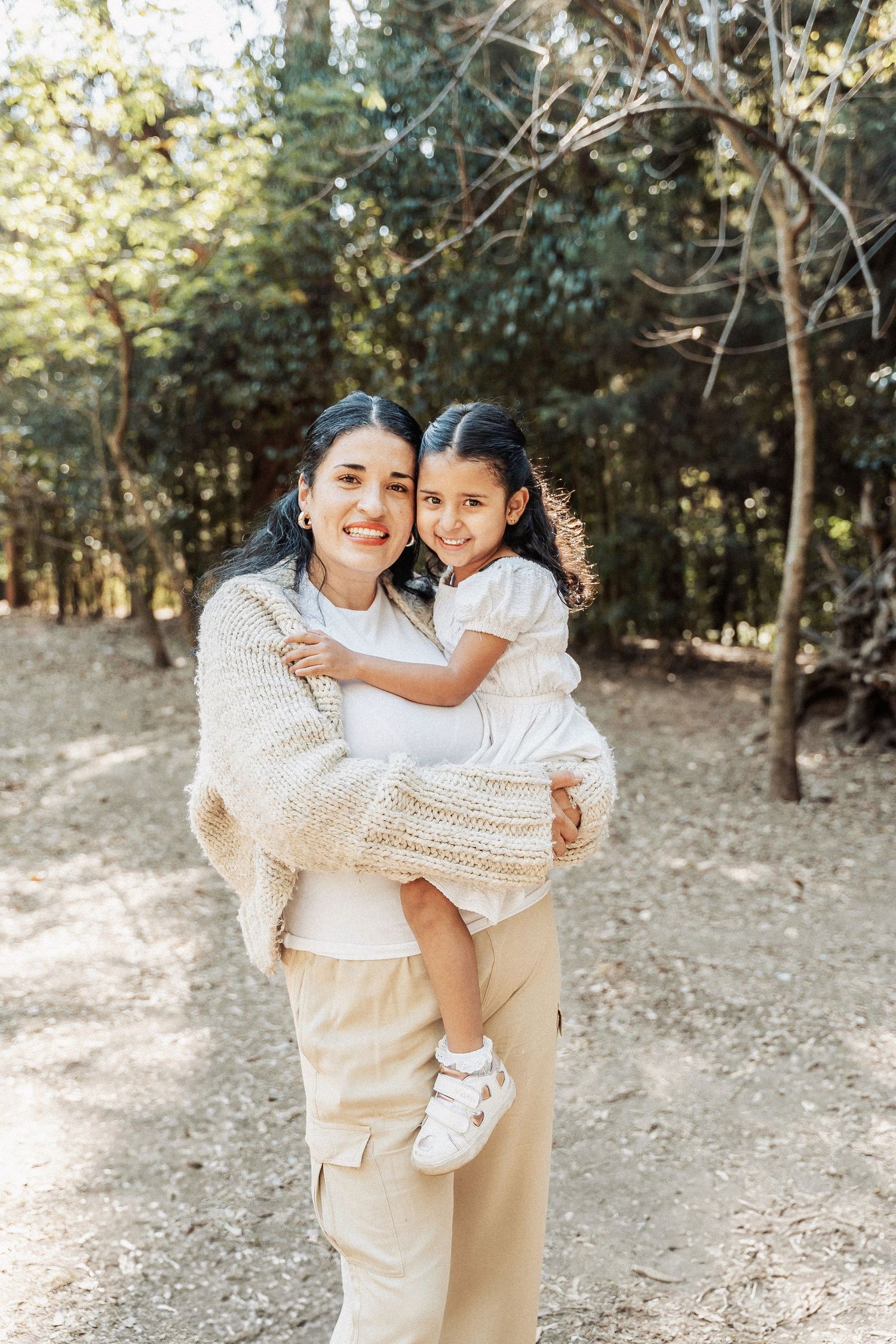 A loving embrace in nature shows two figures wearing white and beige outfits standing in a wooded outdoor setting.