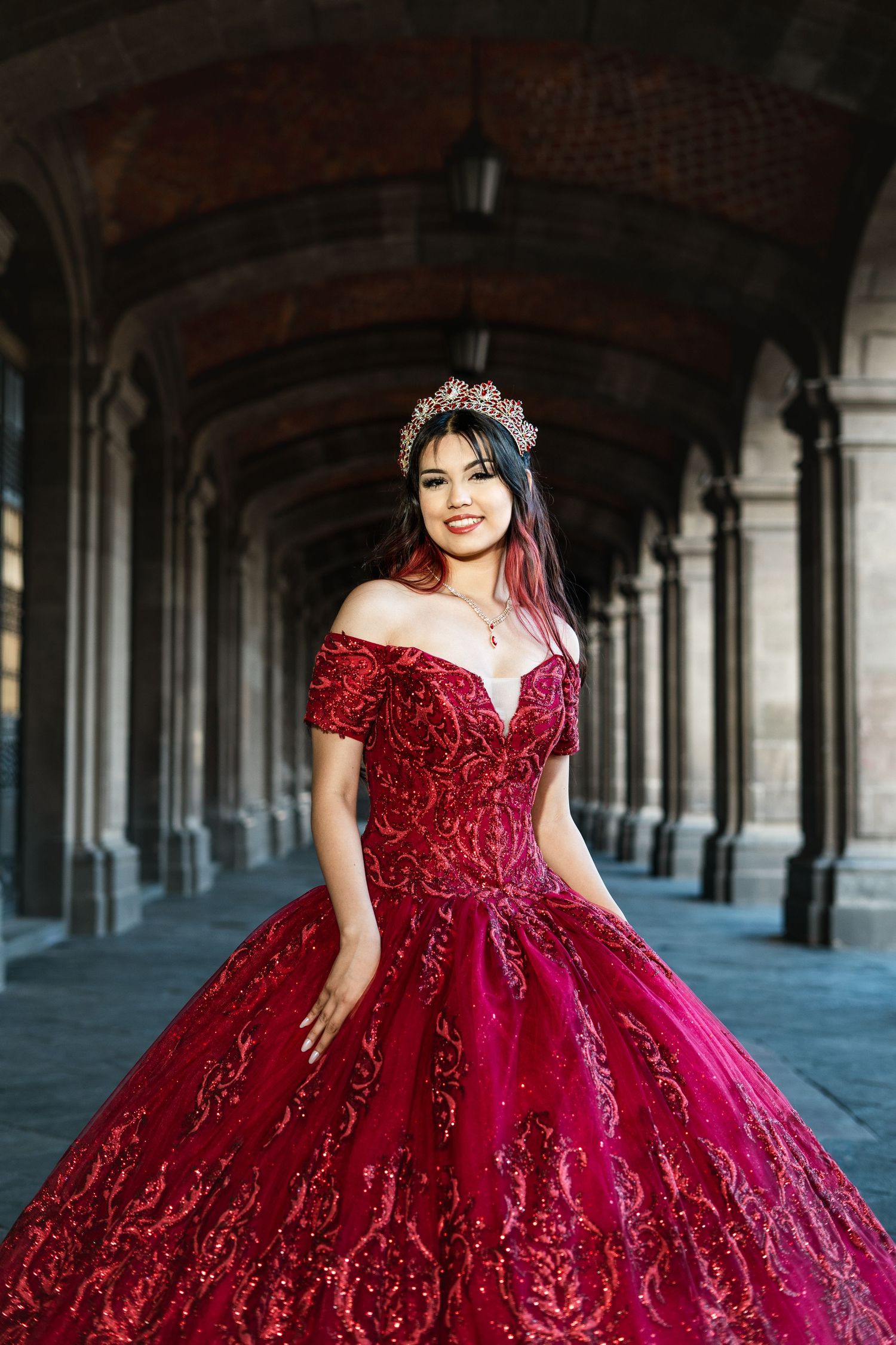 Woman in an ornate burgundy off-shoulder ball gown with beaded details poses in a columned hallway.