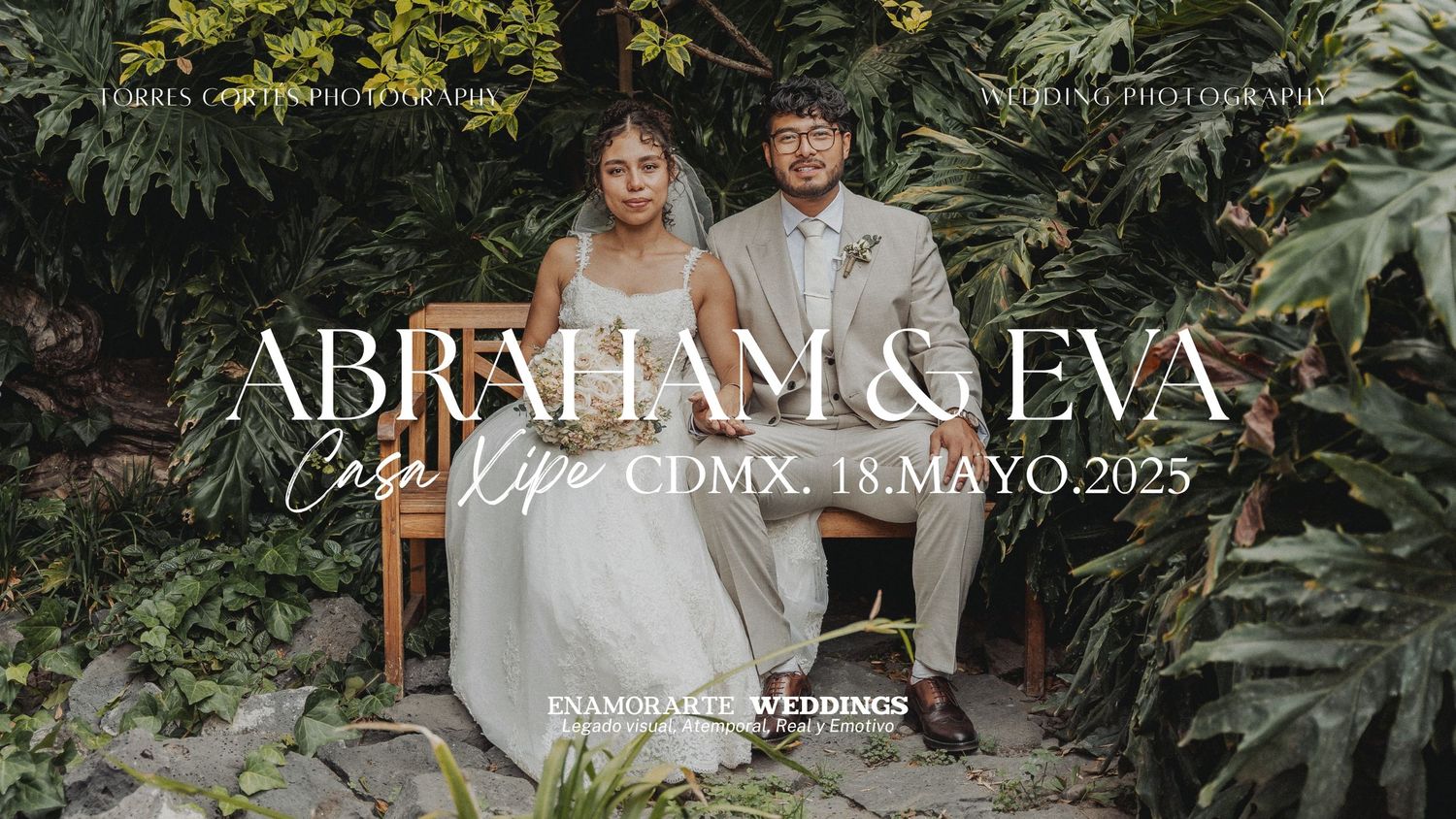 A couple in wedding attire sit on a white bench surrounded by lush tropical greenery.
