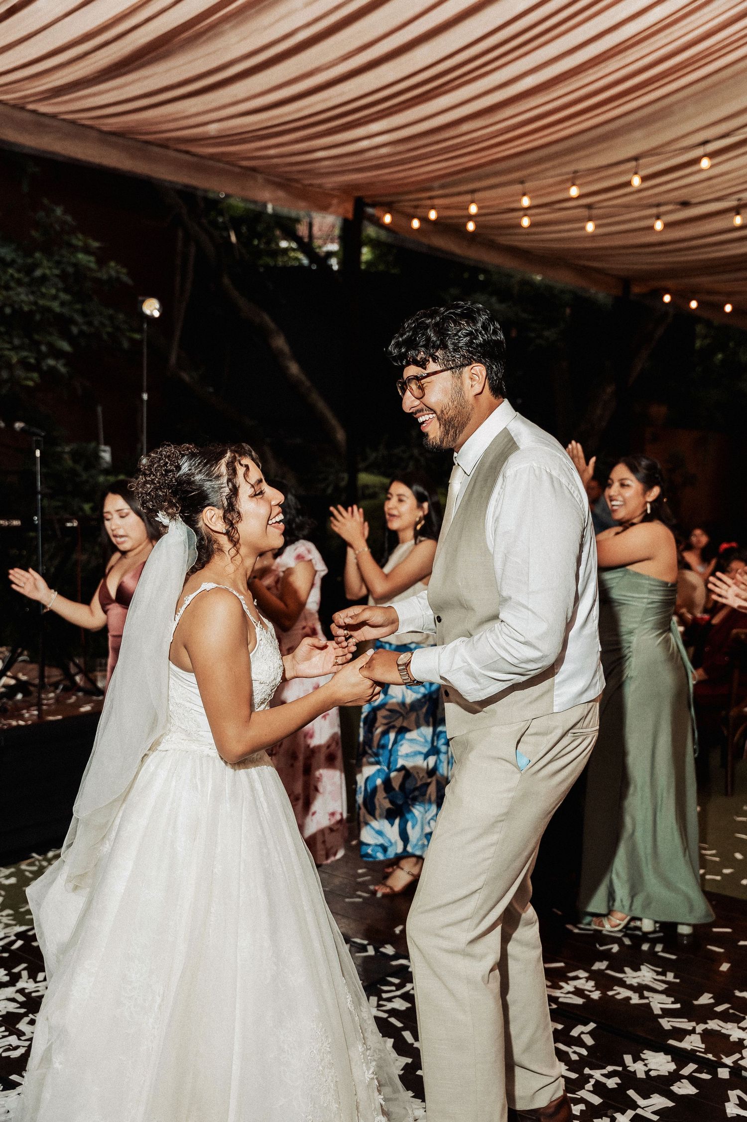 Wedding couple and guests dancing under an elegant fabric-draped canopy with string lights.