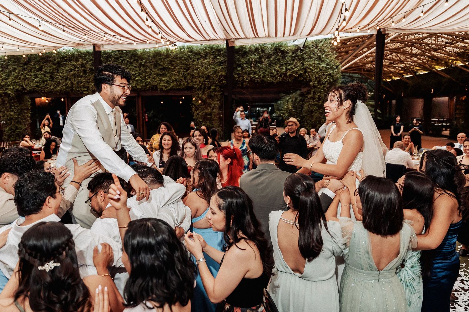Wedding reception guests dance and celebrate while hoisting up newlyweds in chairs during the Hora dance tradition under a tent.