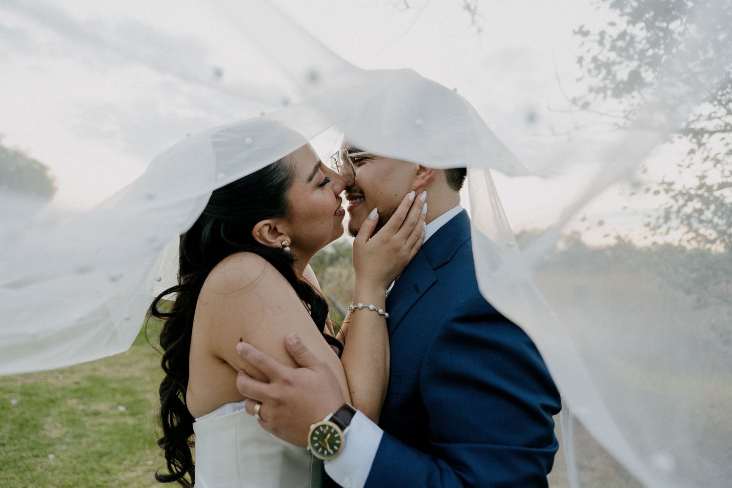 A couple shares a romantic kiss under a flowing veil during an outdoor wedding ceremony.