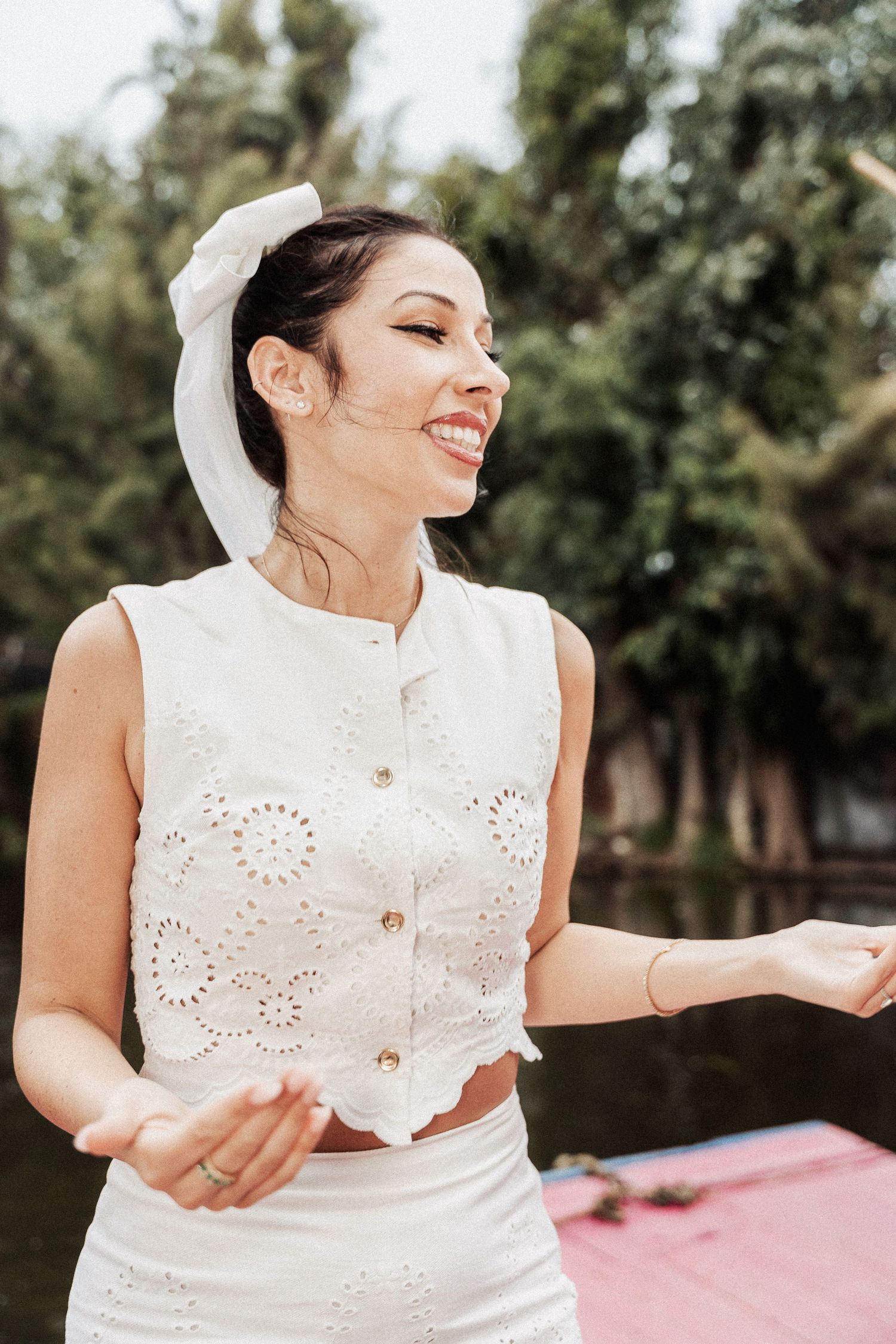 A woman in a white eyelet crop top smiles joyfully while standing outdoors in natural lighting.