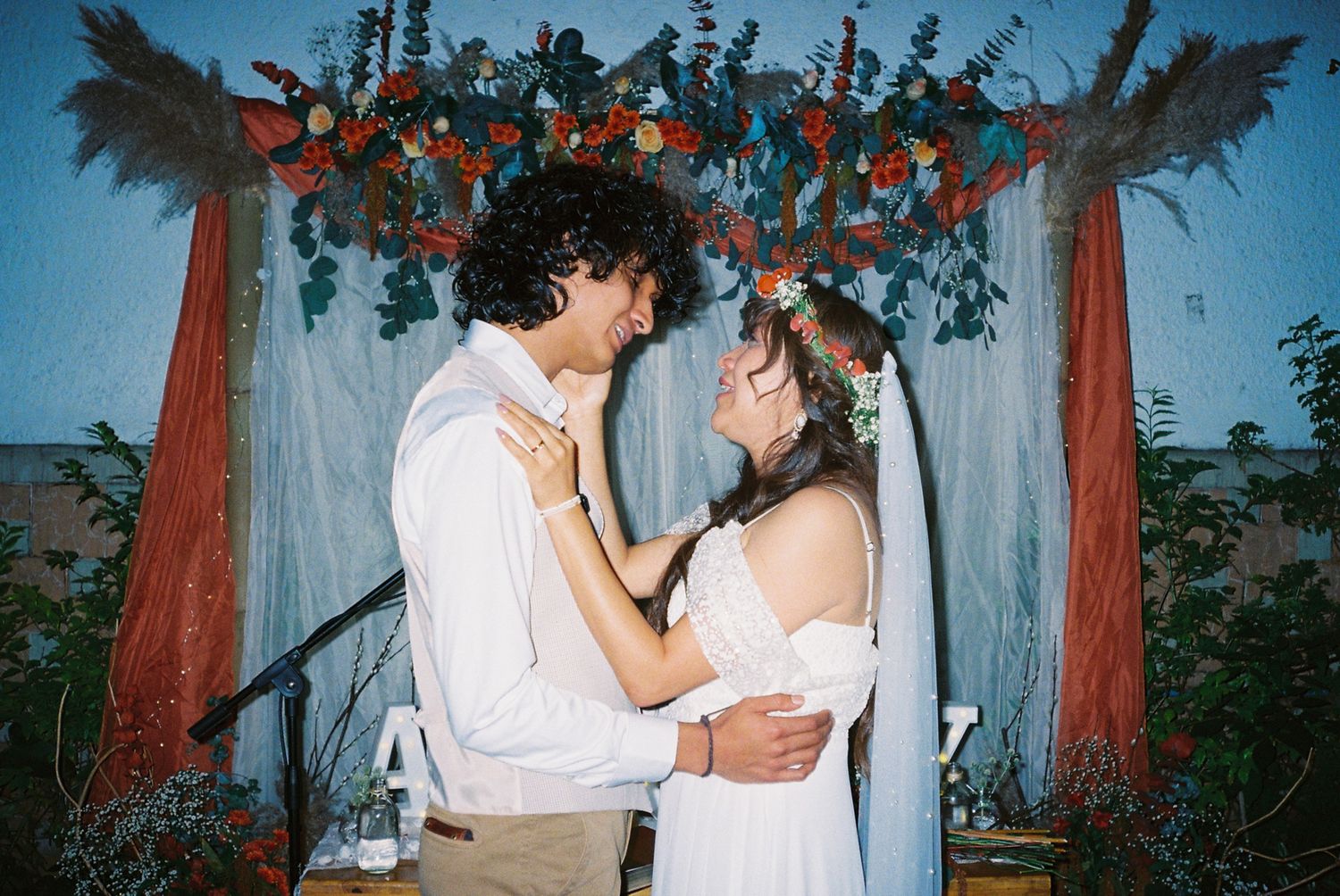A romantic couple shares an intimate moment under a draped outdoor canopy with blue lighting and trees in the background.