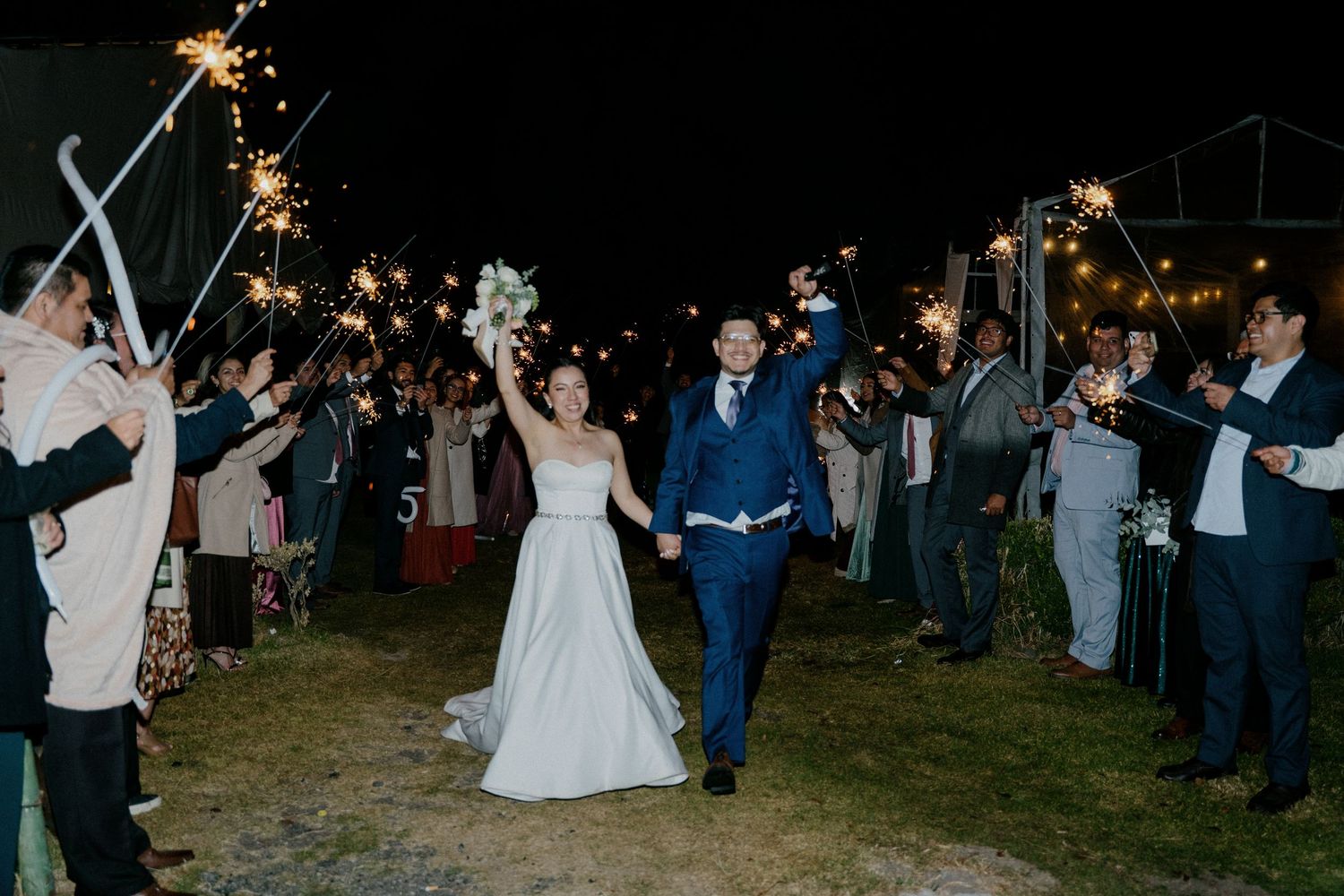 Wedding guests hold sparklers creating a magical nighttime send-off tunnel for the newlyweds.