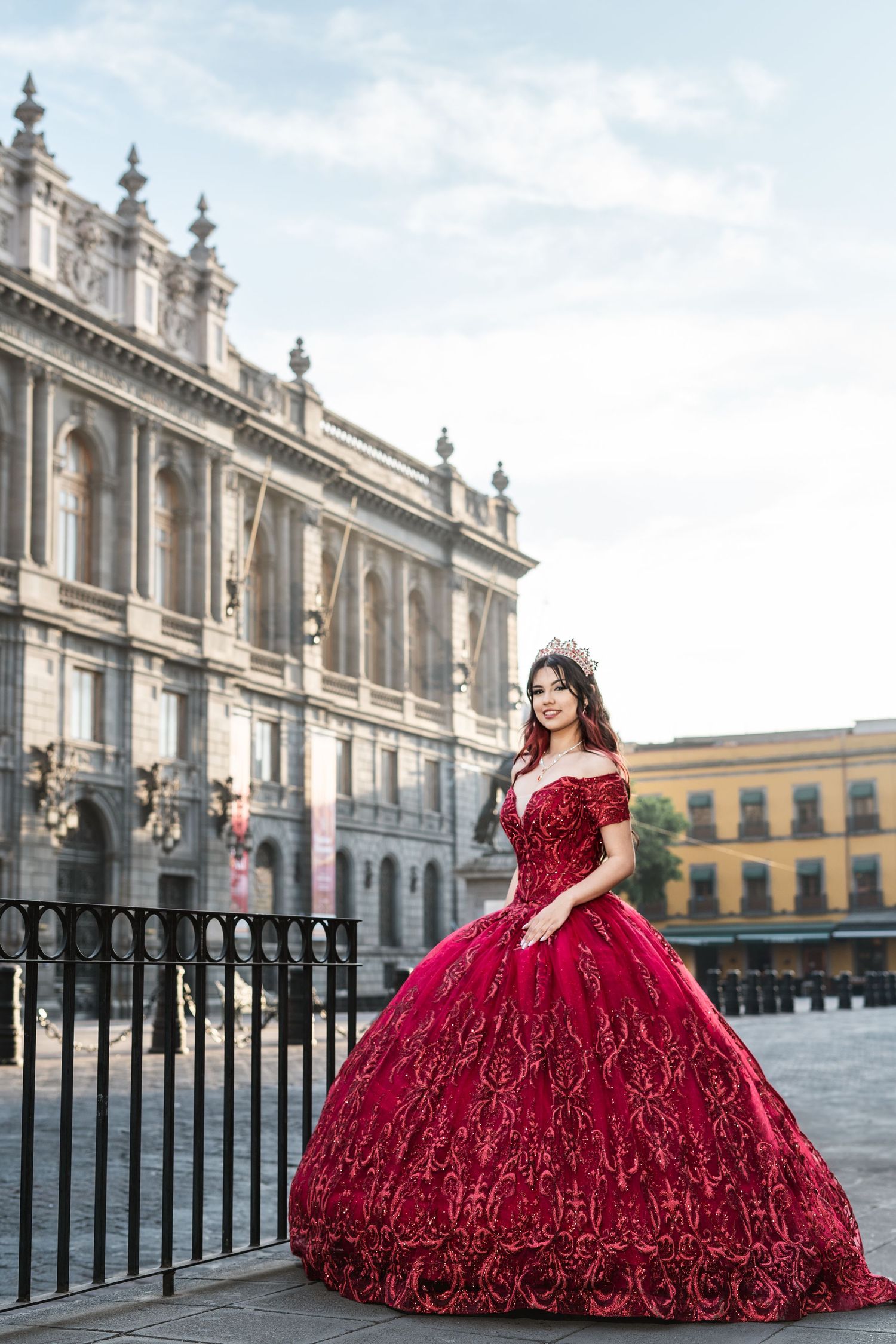 Stunning red ball gown with off-shoulder design photographed against ornate historic European architecture background.