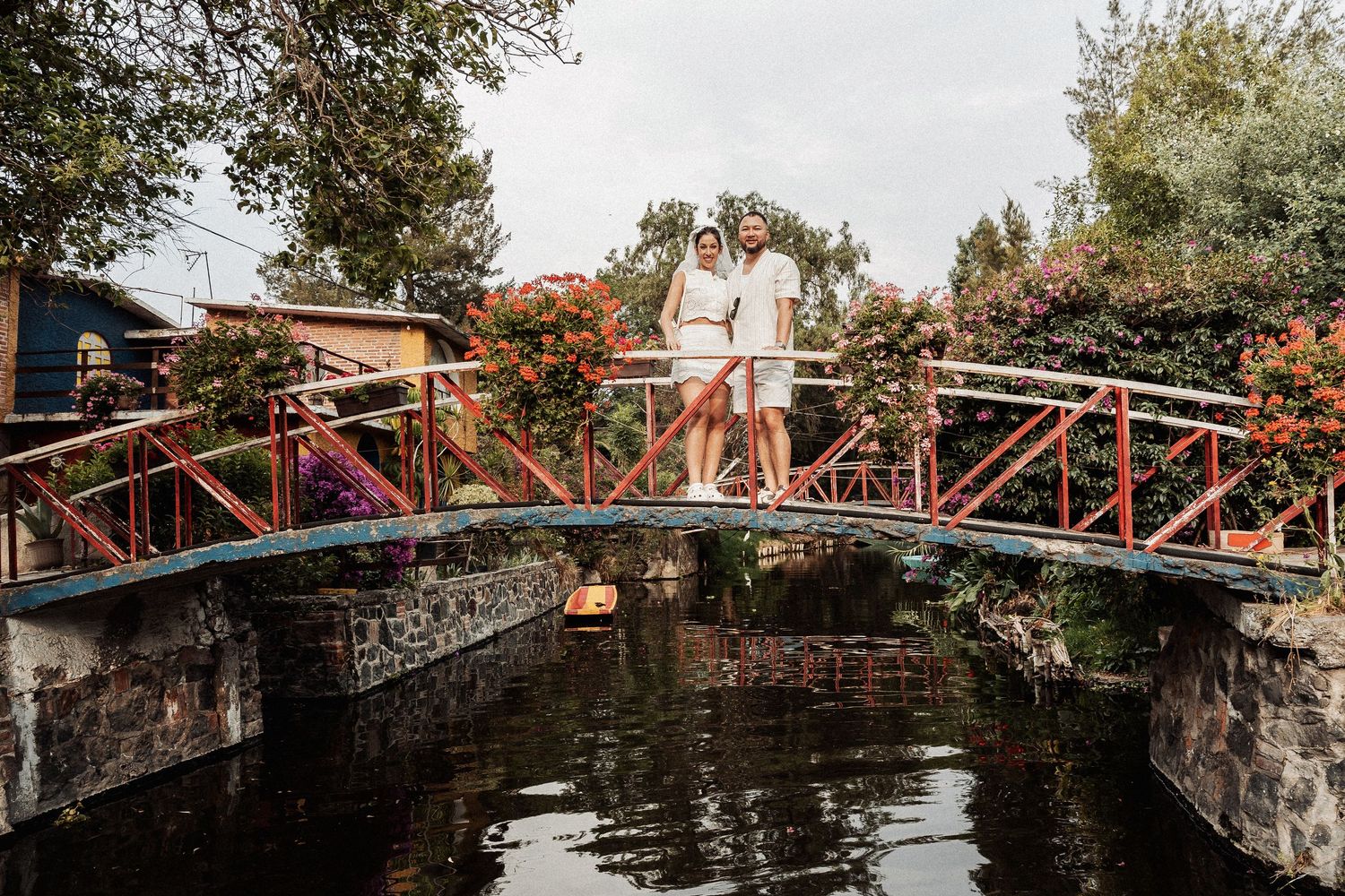A couple stands on a rustic red iron footbridge over a stone-lined canal surrounded by autumn foliage.