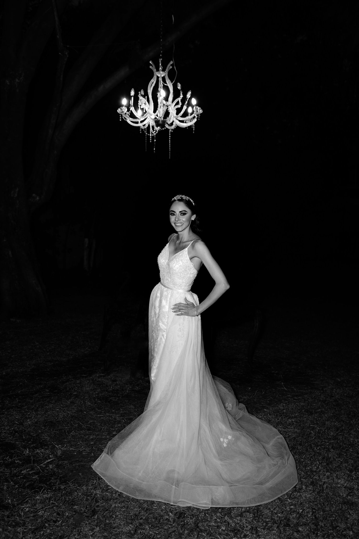 A bride in a flowing white gown poses under an ornate chandelier in a dramatic black and white photo.