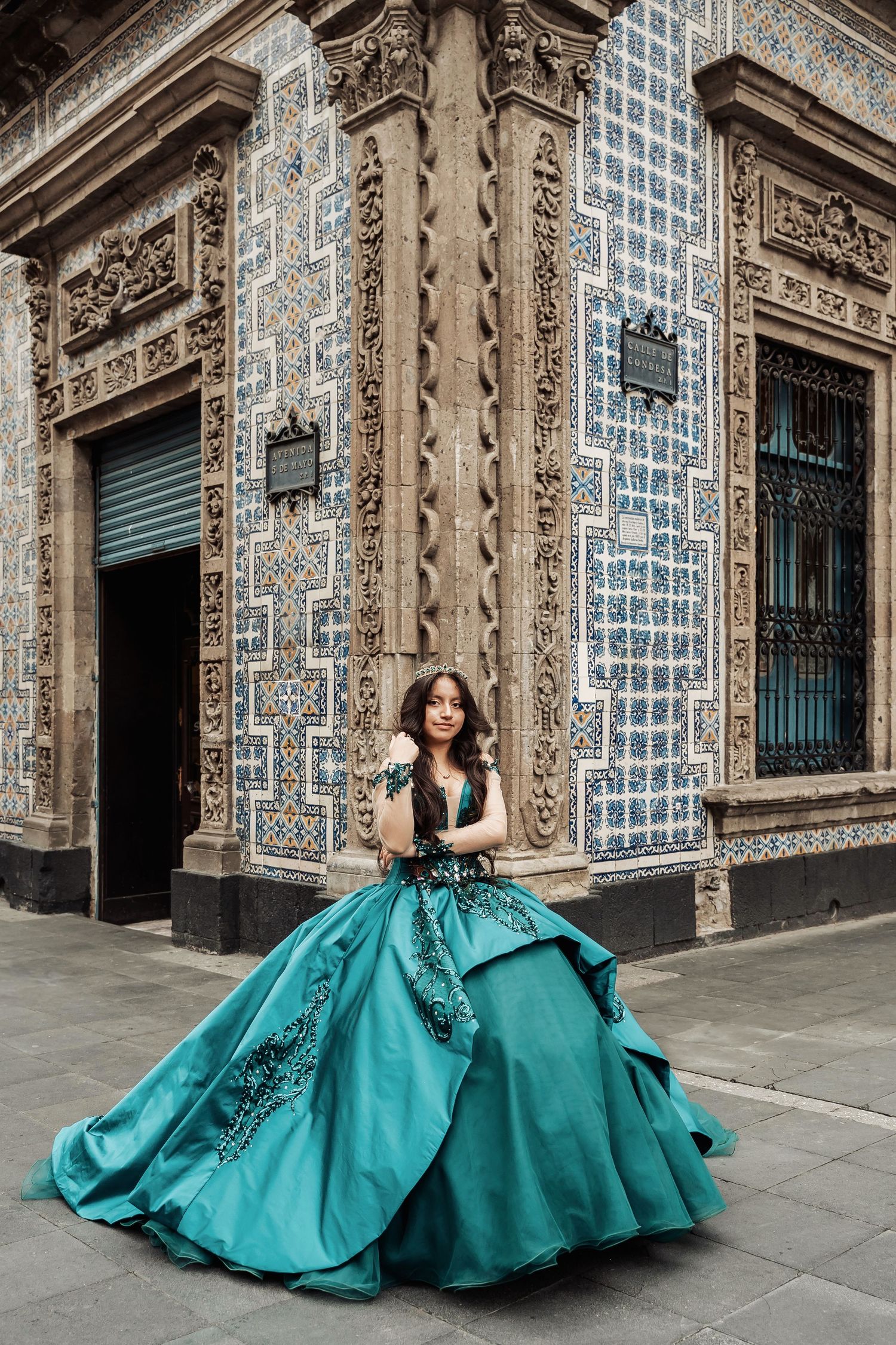 A person wearing an elegant turquoise ballgown poses gracefully against an ornate tiled colonial building exterior in Mexico.