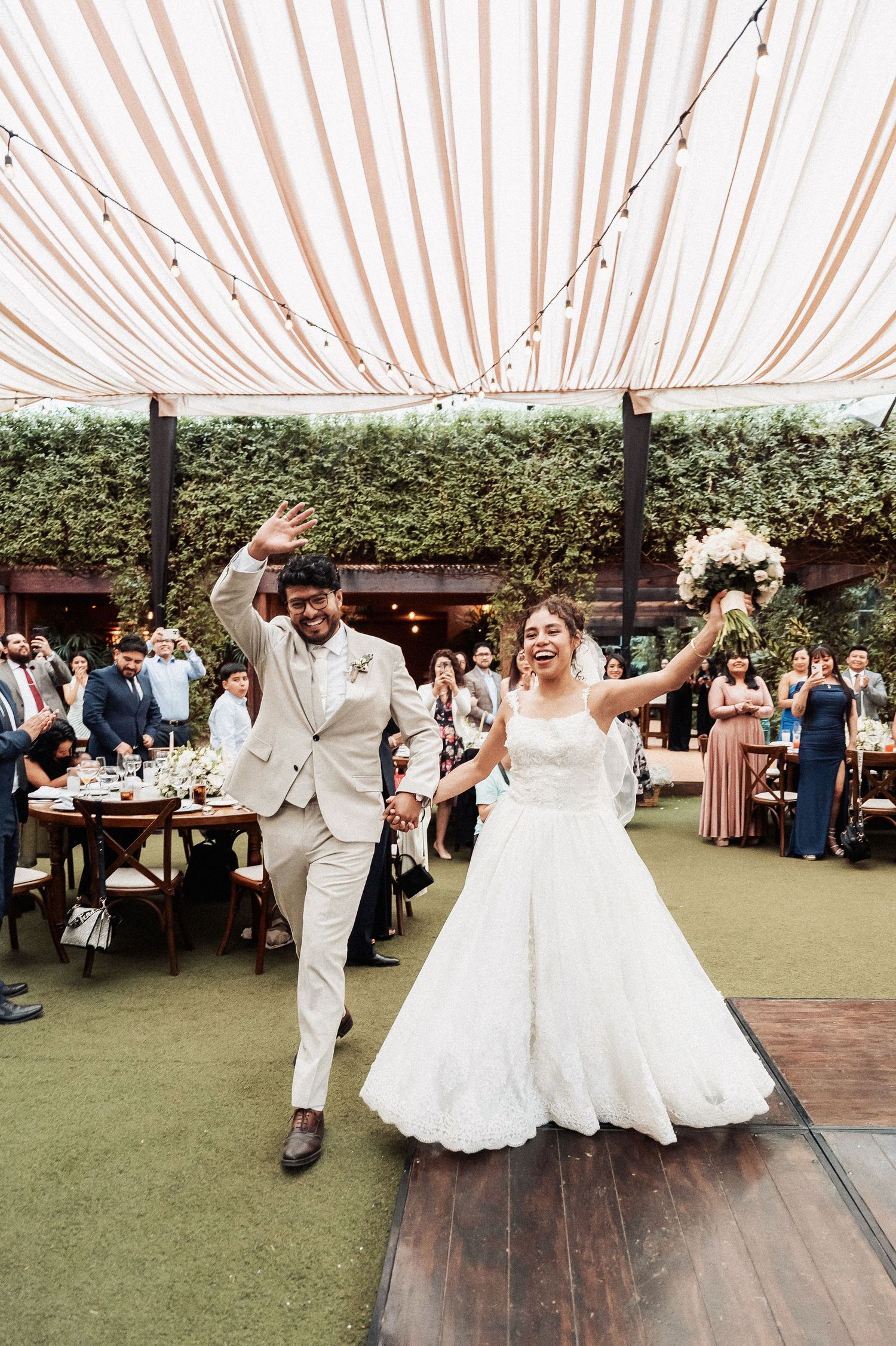 Newlyweds and wedding party dancing energetically under a white tent at an outdoor reception celebration.