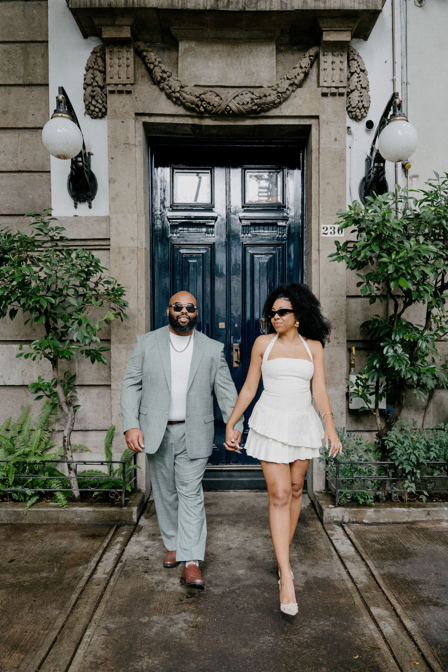 A stylish couple in sunglasses walking together in front of an ornate blue door with decorative stone archway.