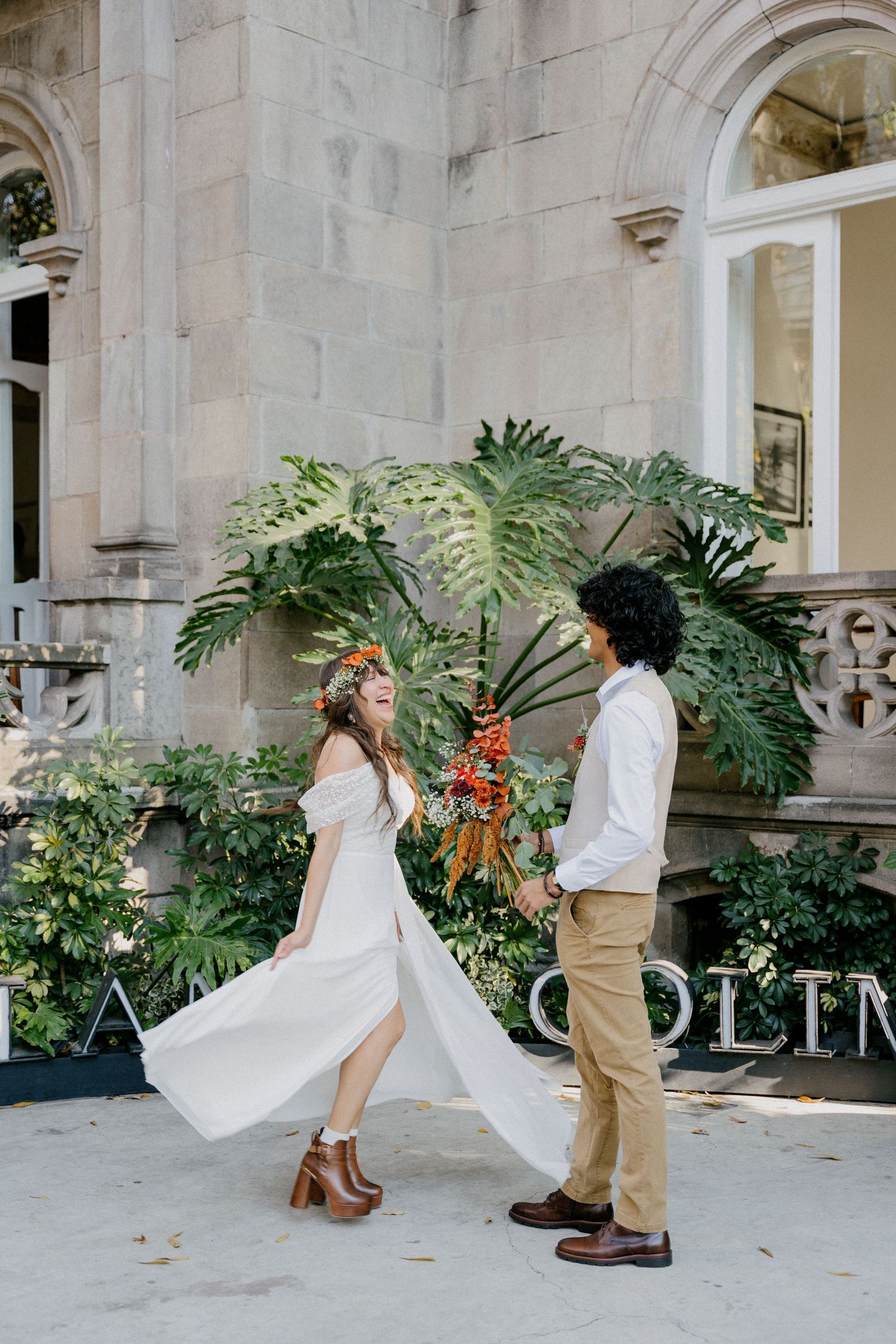 A couple in wedding attire dance together on a stone patio with lush tropical plants and architecture in the background.