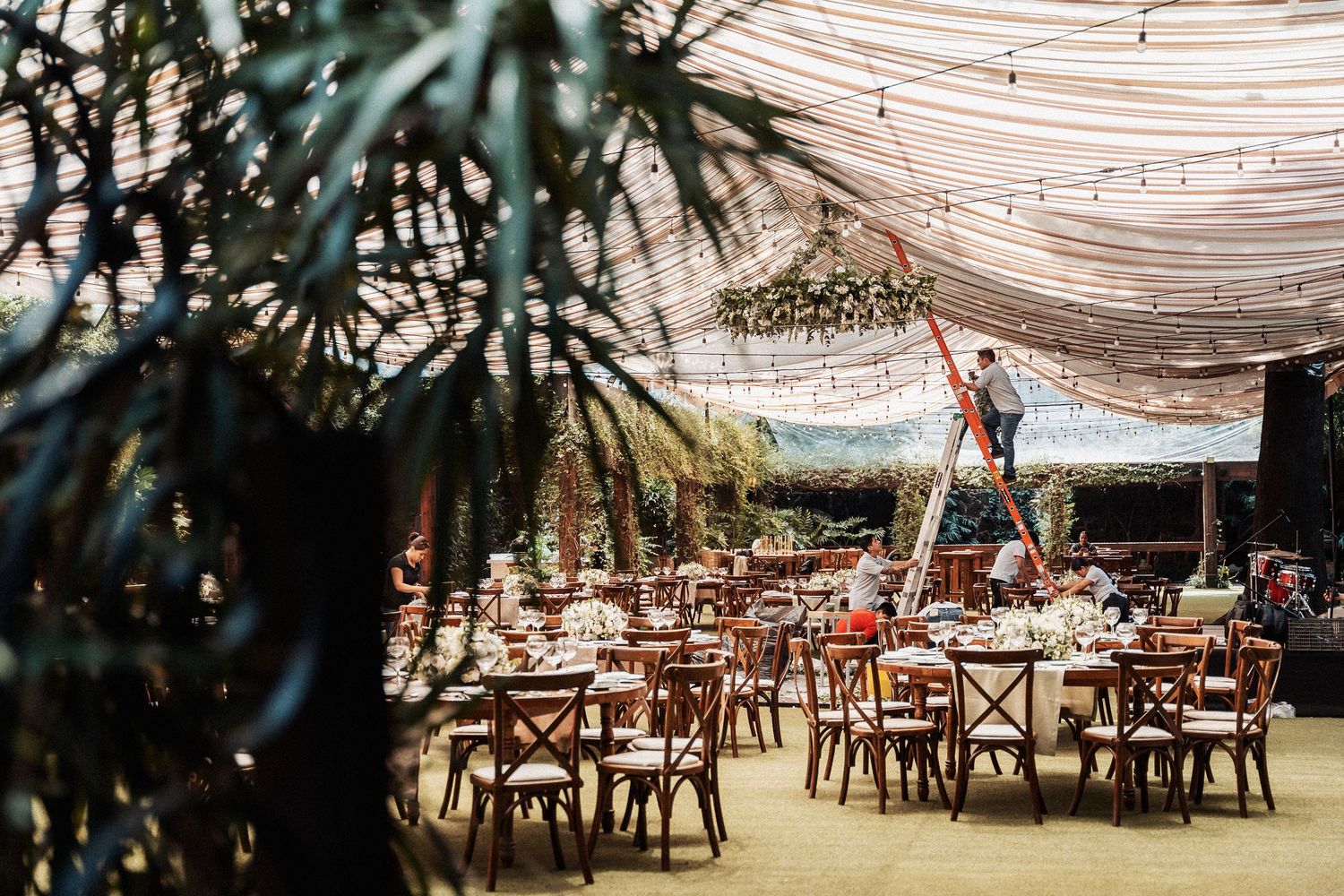 String lights illuminate an outdoor dining area with rustic wooden tables and chairs under a draped canopy at sunset.
