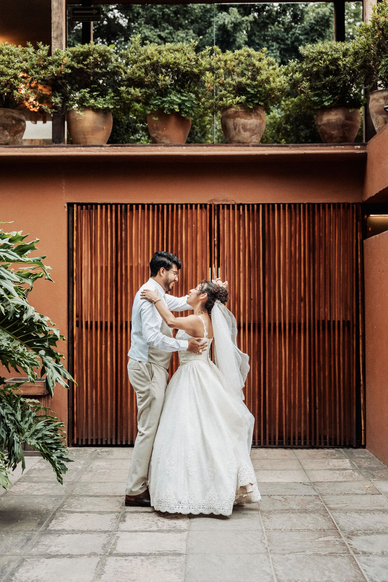A couple in wedding attire share an intimate dance moment in front of a wooden slatted door and terra cotta wall.