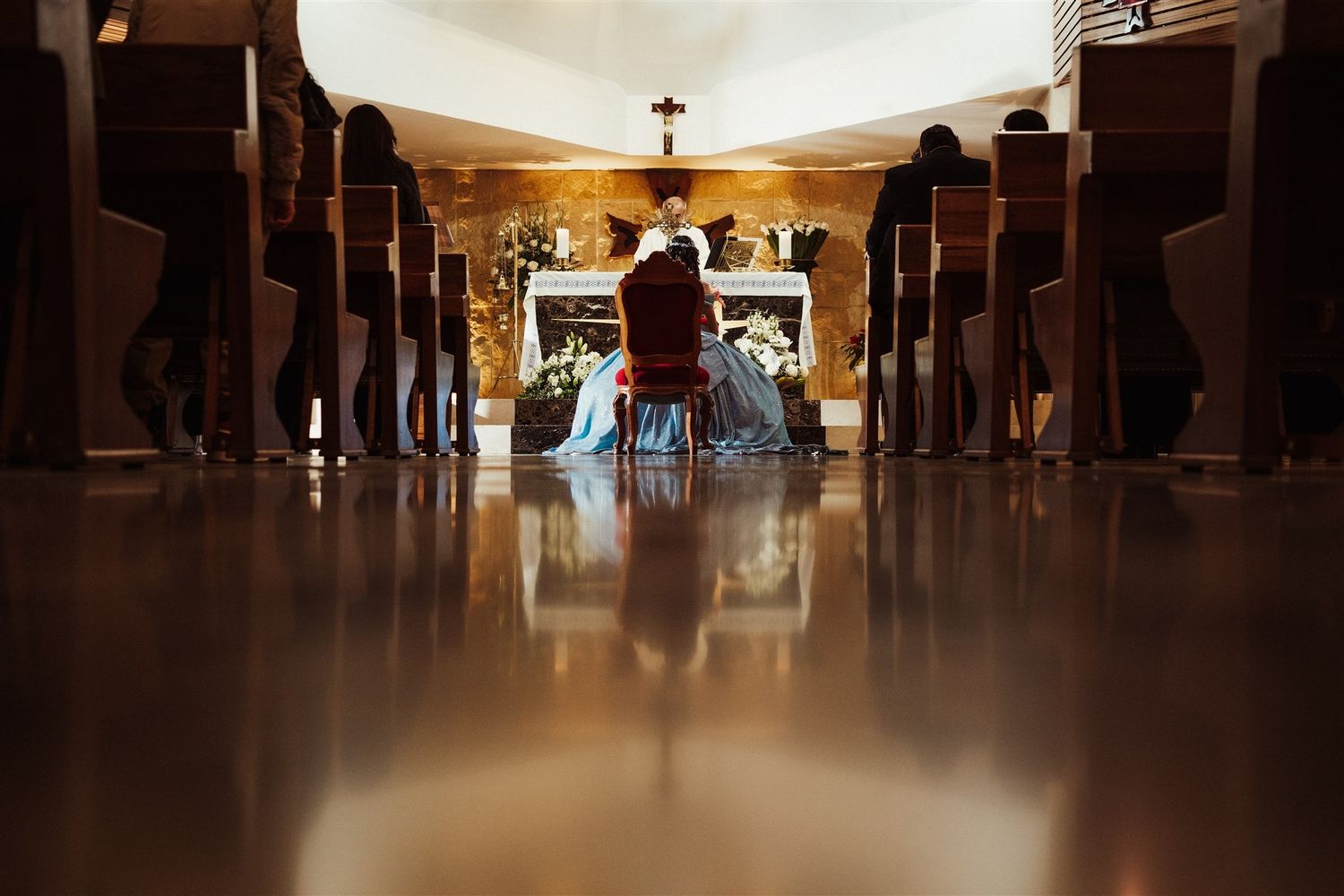 Interior view down the center aisle of a church with wooden pews and warm lighting casting reflections on the floor.