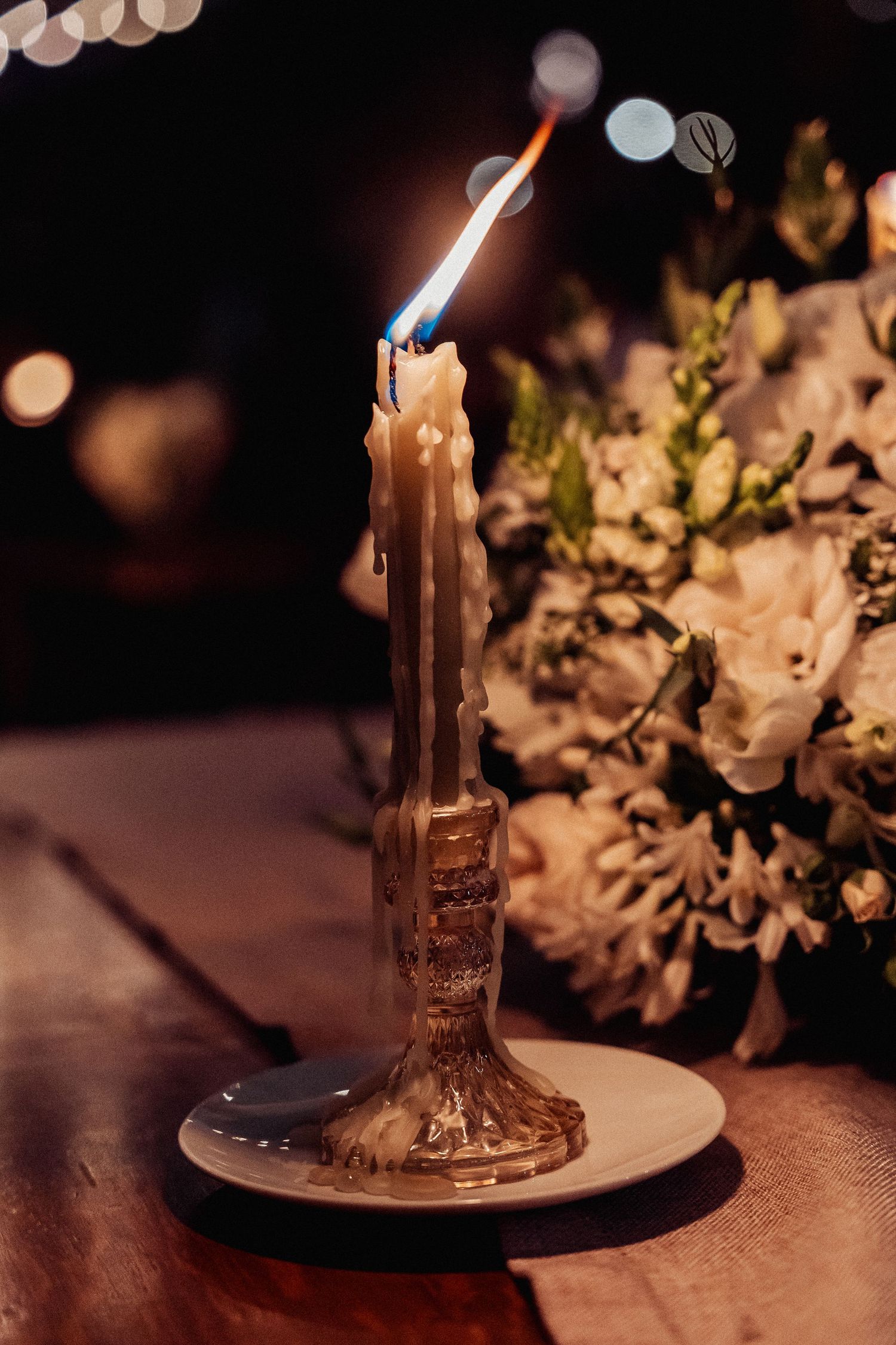 A melting candle flickers on a plate surrounded by soft focus white flowers in a moody, romantic setting.