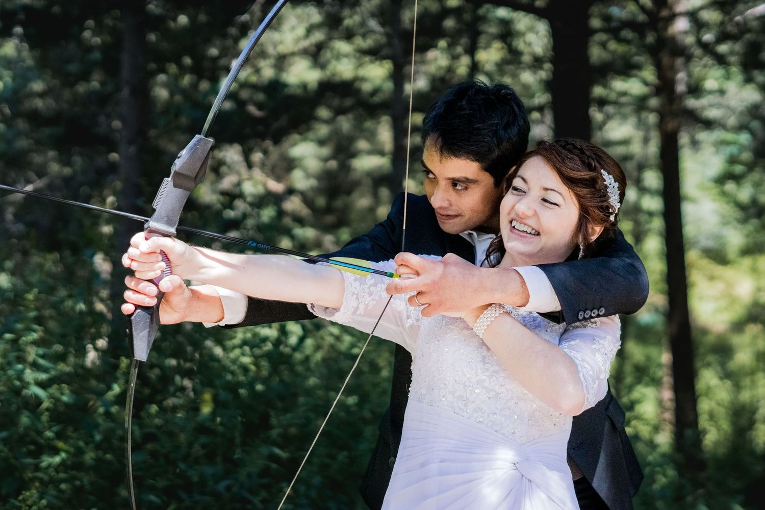 A couple practices archery together in a forest setting during their wedding photoshoot.