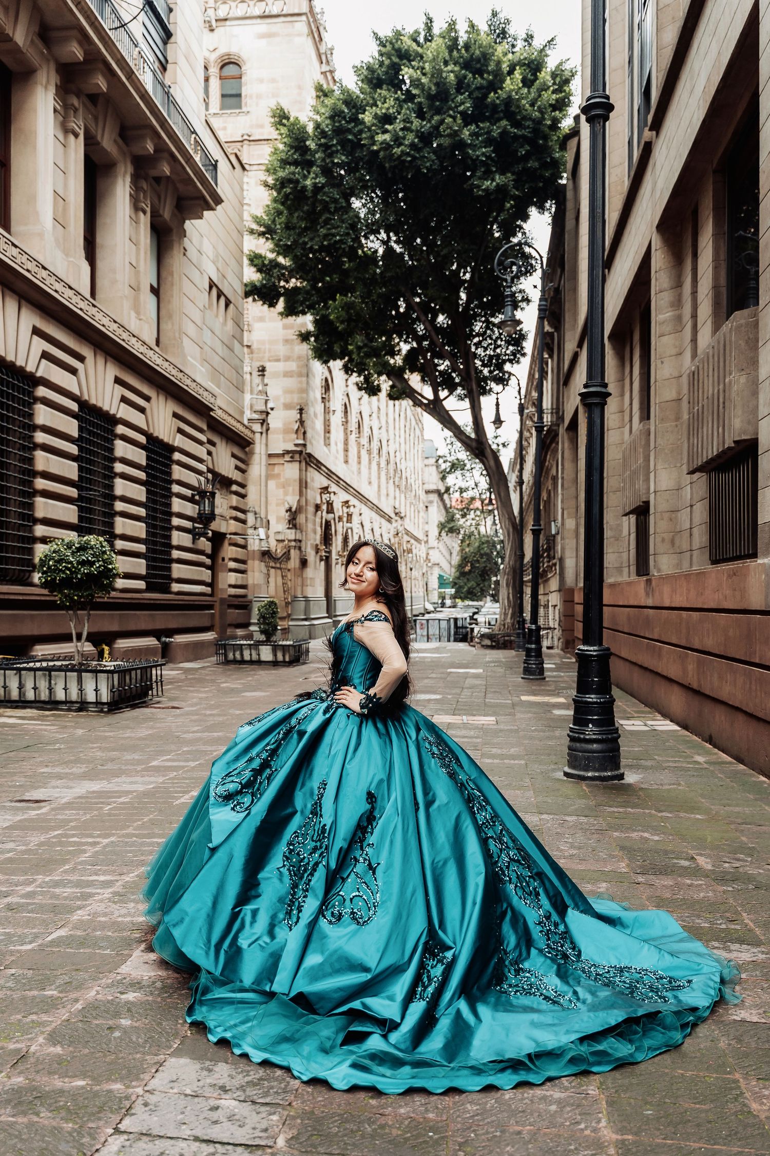 Elegant teal ballgown with black lace details sweeps across cobblestone street between historic buildings.