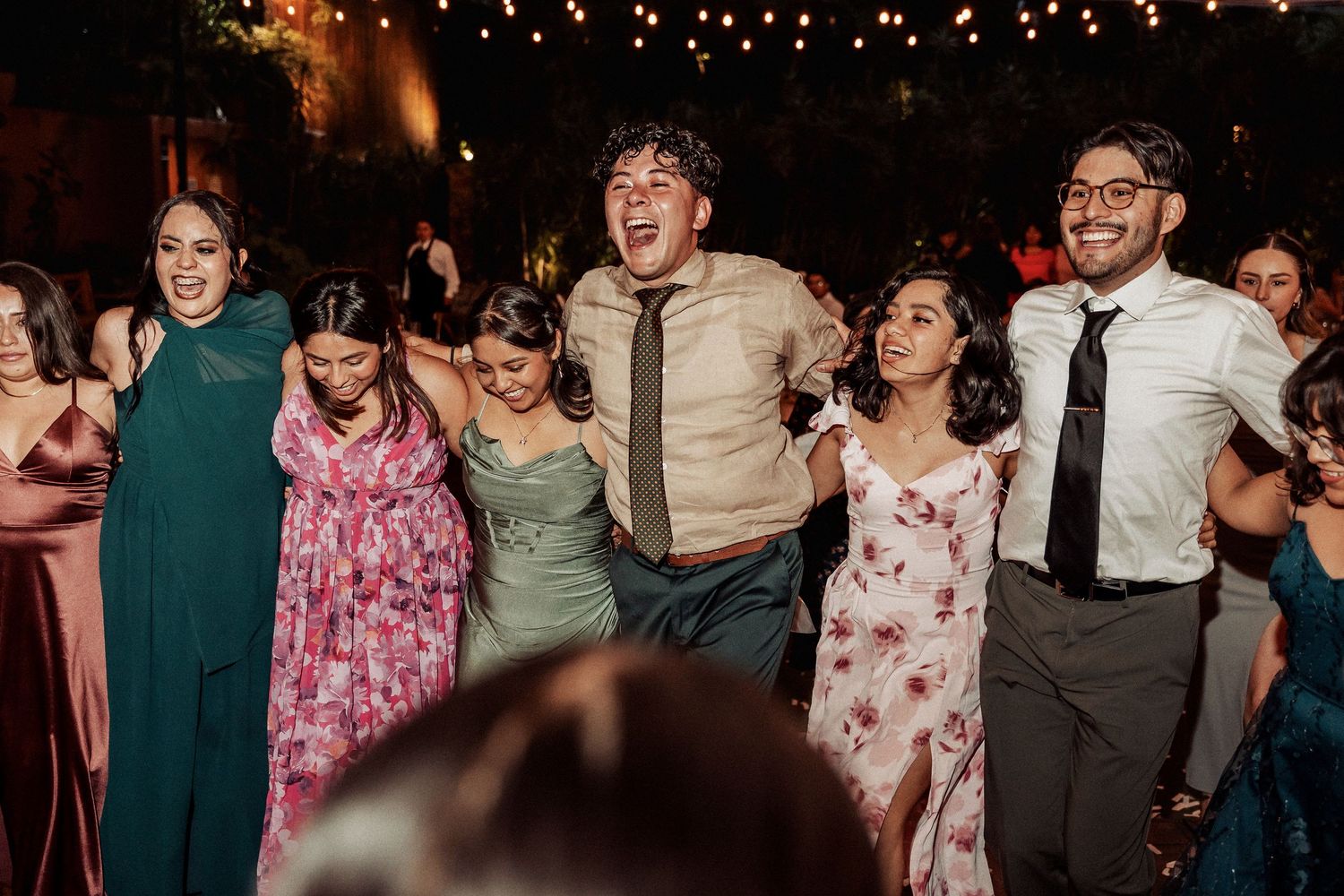 A group of wedding guests dance and laugh together on the dance floor under string lights at an evening reception.