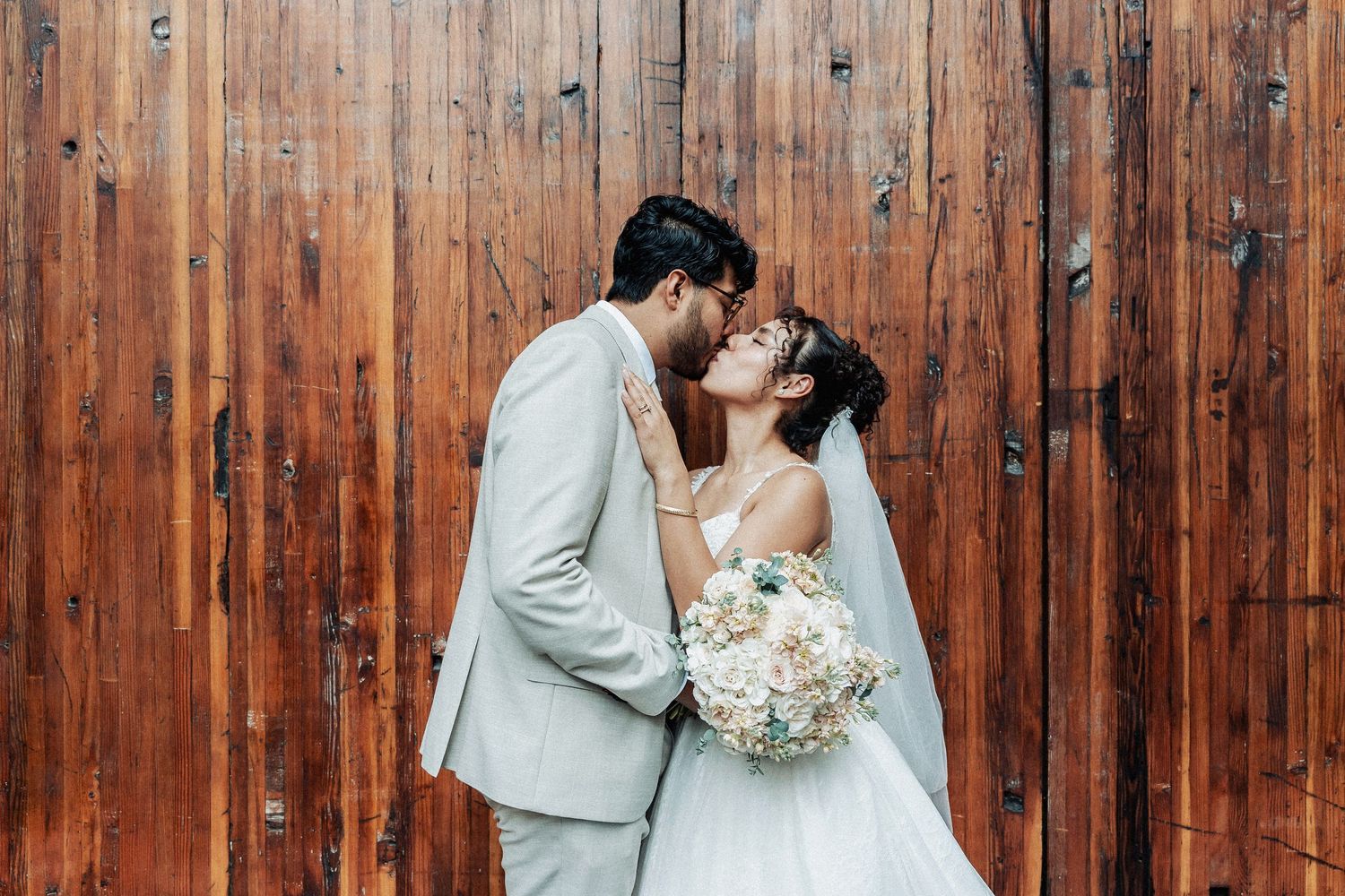 A romantic newlywed couple embraces against a rustic wooden wall backdrop, with the bride holding a soft white bouquet.