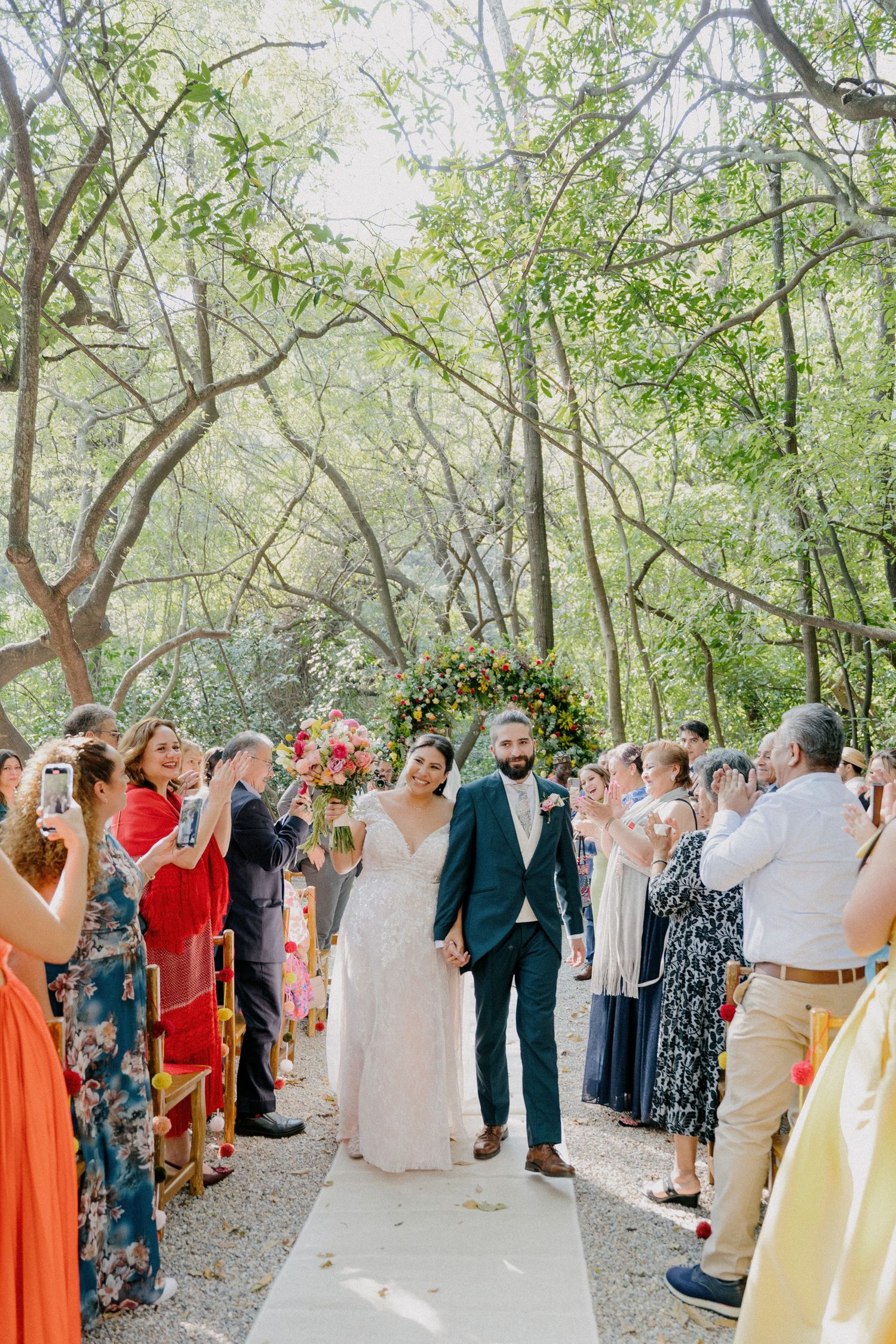 Wedding guests gather outdoors under a canopy of trees during a daytime celebration ceremony.