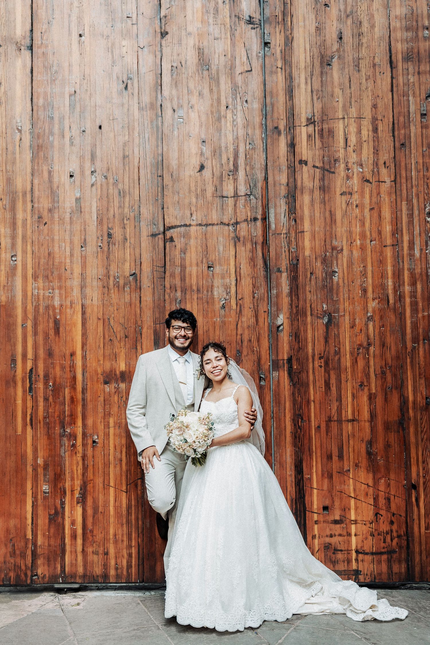 Wedding couple poses against rustic wooden wall background in formal attire with white dress and beige suit.