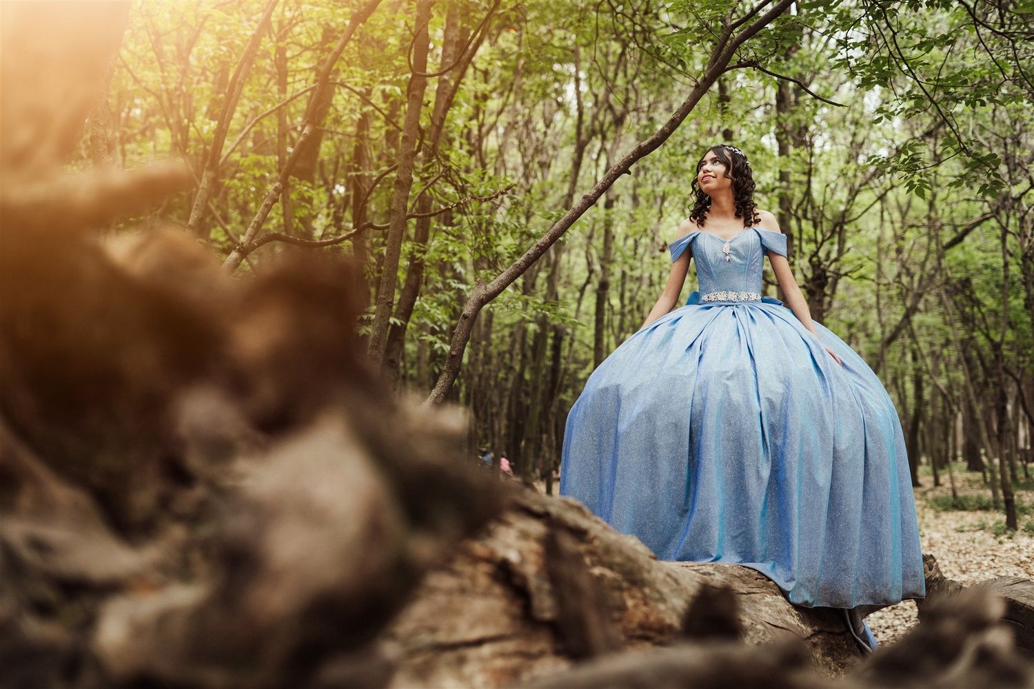 A person in a flowing blue ballgown dress walks through a sunlit woodland path creating a magical atmosphere in nature.