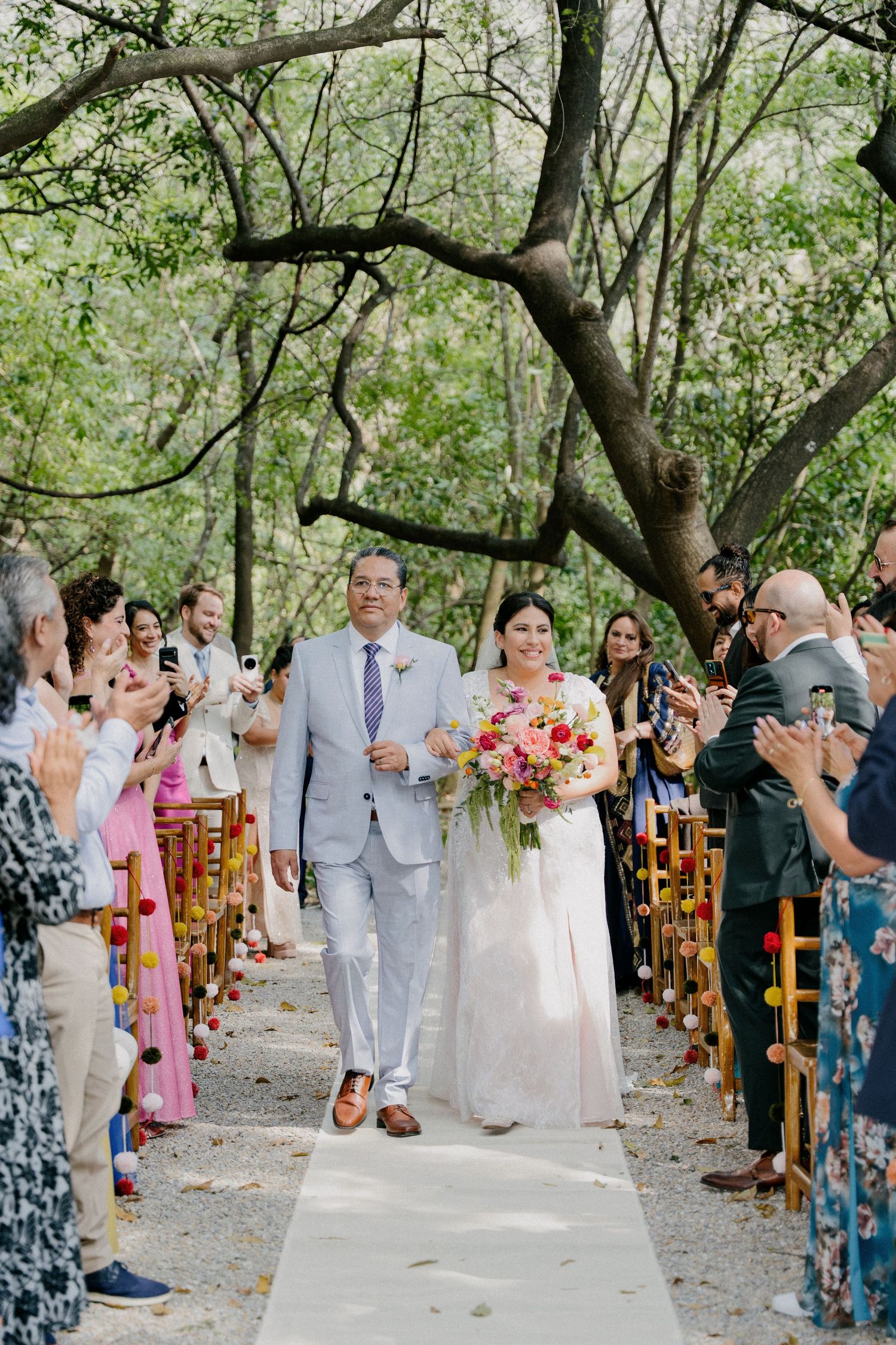 Wedding guests line a garden path taking photos of the newlyweds during an outdoor summer ceremony.