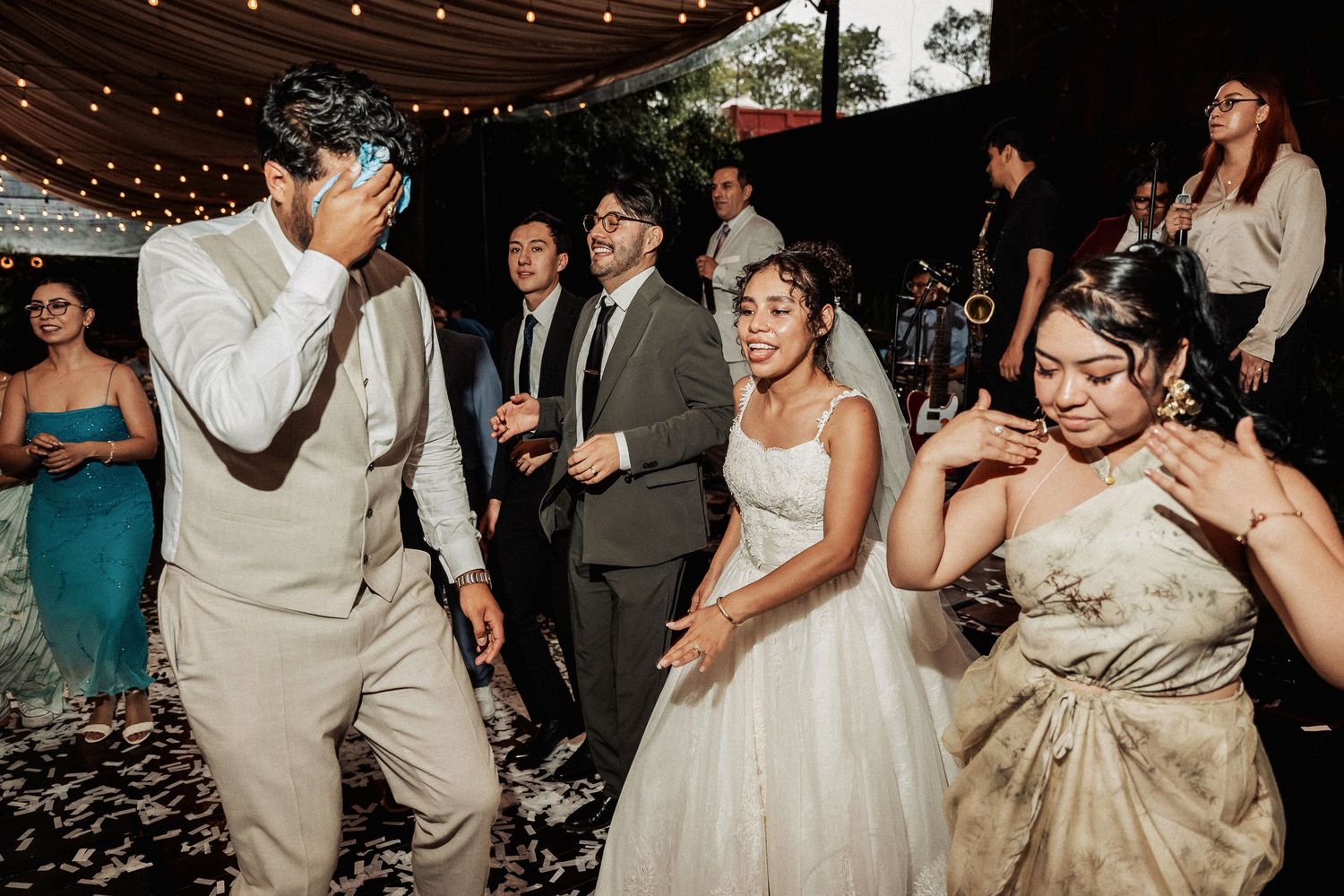 Wedding party dancing under string lights and fabric draping at outdoor evening celebration.