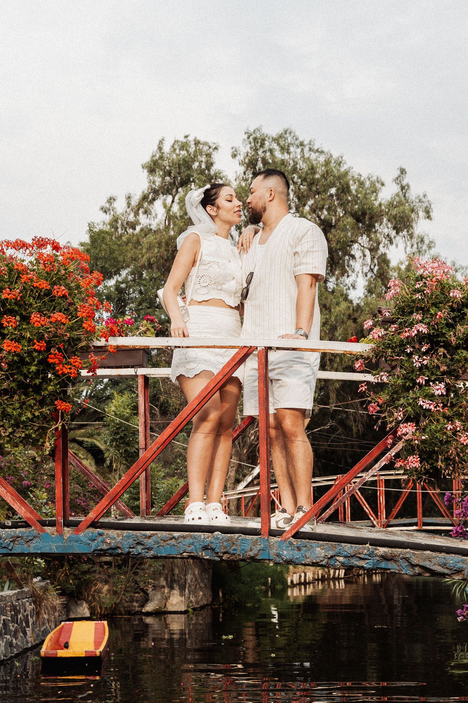 Couple in white outfits standing together on a red metal bridge surrounded by blooming red flowers near water.