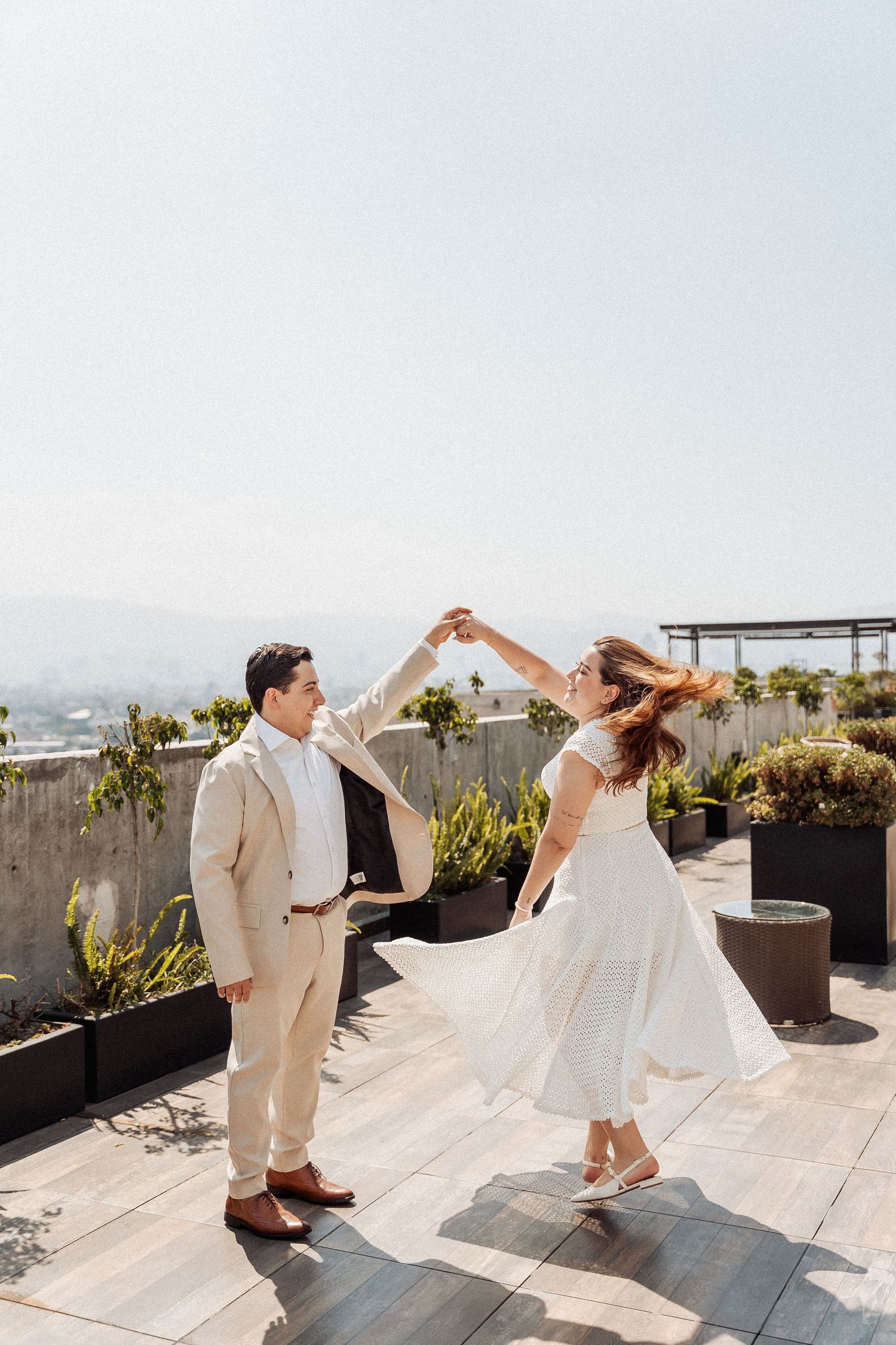 A couple dance together on a rooftop terrace with plants and cityscape views in the background.