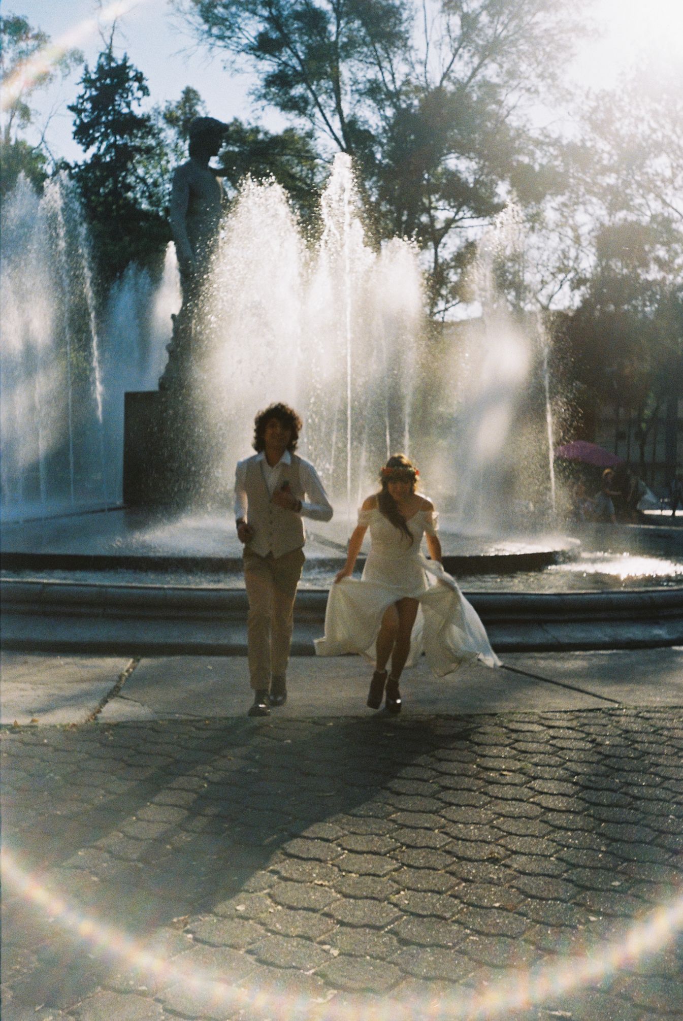 People silhouetted against sunlit water fountains in a park with stone paving and dramatic backlit spray.